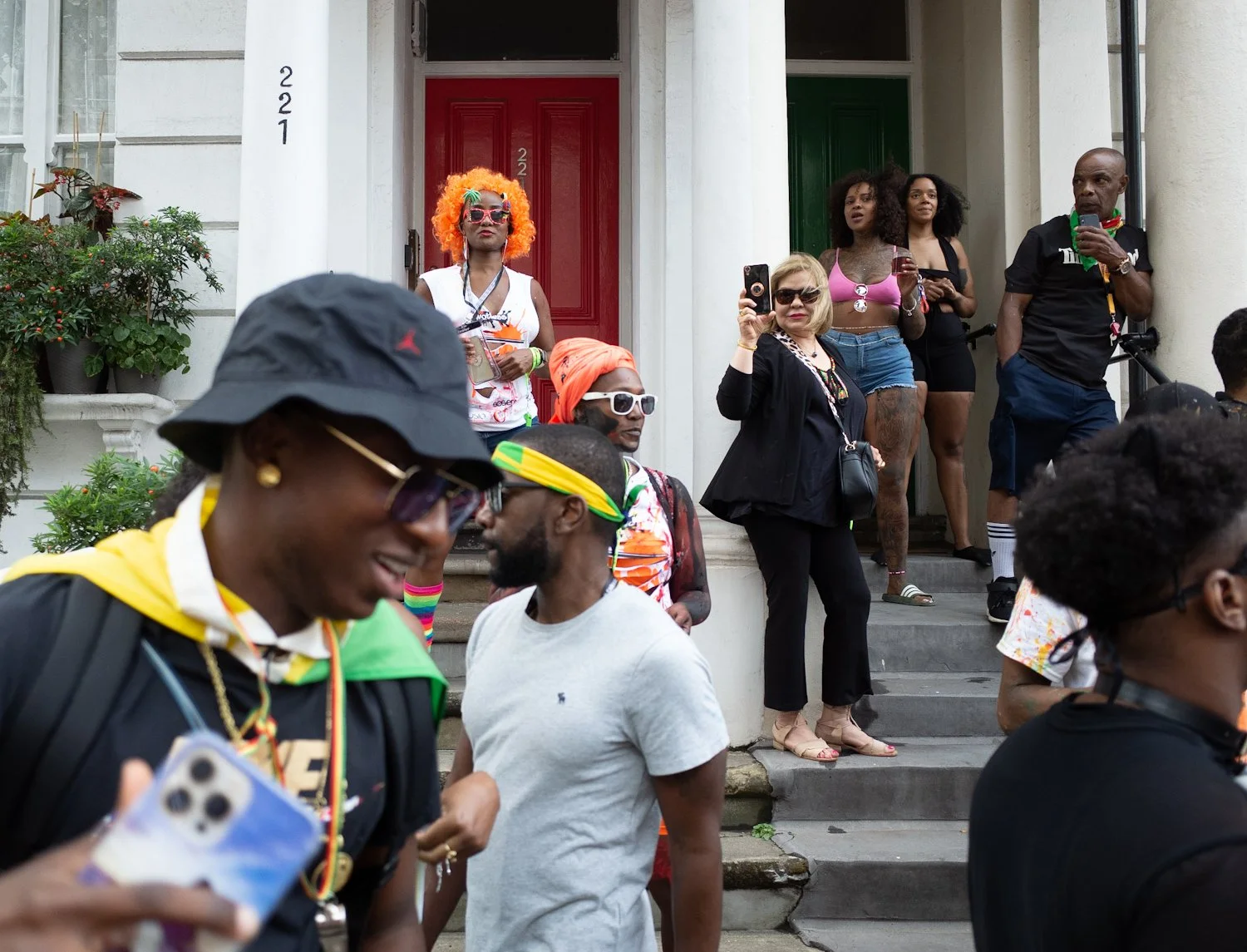 People gathered on the steps of a building, some taking photos or videos, with diverse clothing and accessories, celebrating or attending an event.