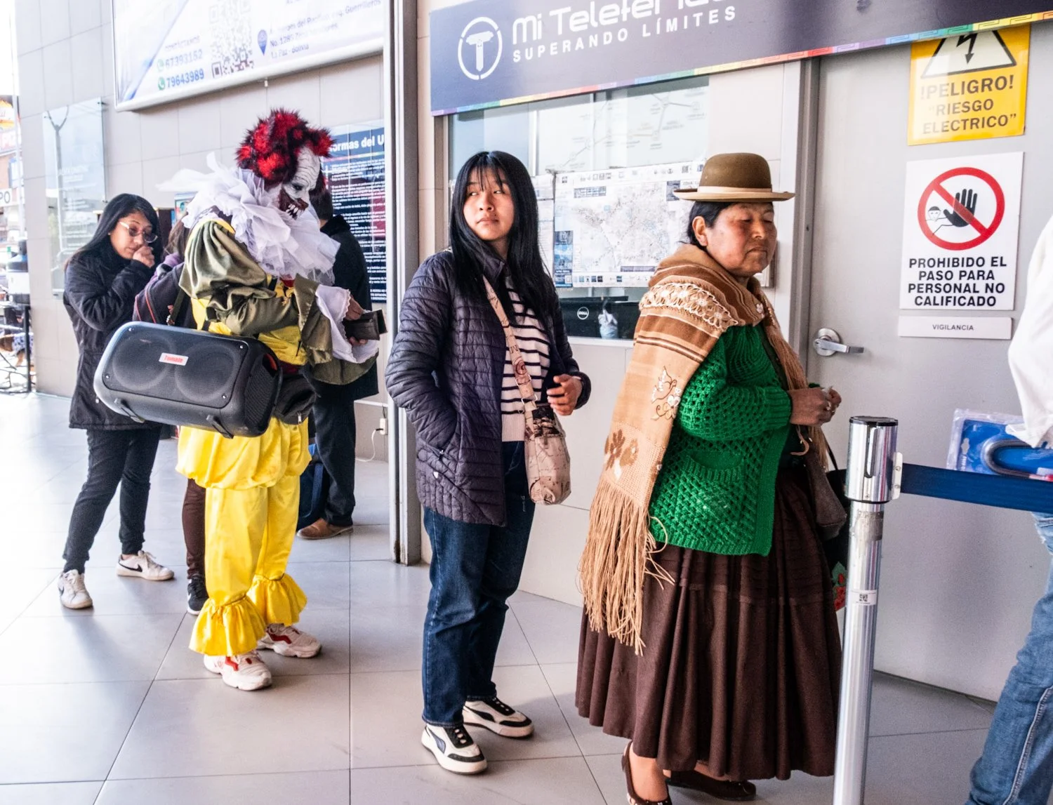 People standing in line at a transportation ticket counter. One person is dressed as Pennywise from It, in a clown costume and red wig. Others are dressed in casual and traditional clothing, waiting for service.