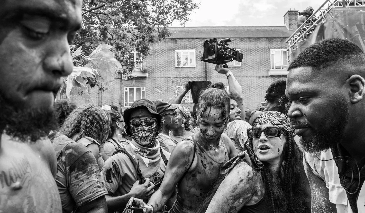 Black and white photo of a group of people outdoors, some covered in dirt and soot, with serious expressions, in a crowded scene. There is a woman in sunglasses with a skull mask and a man with a bun hairstyle in the foreground. A person holds a came