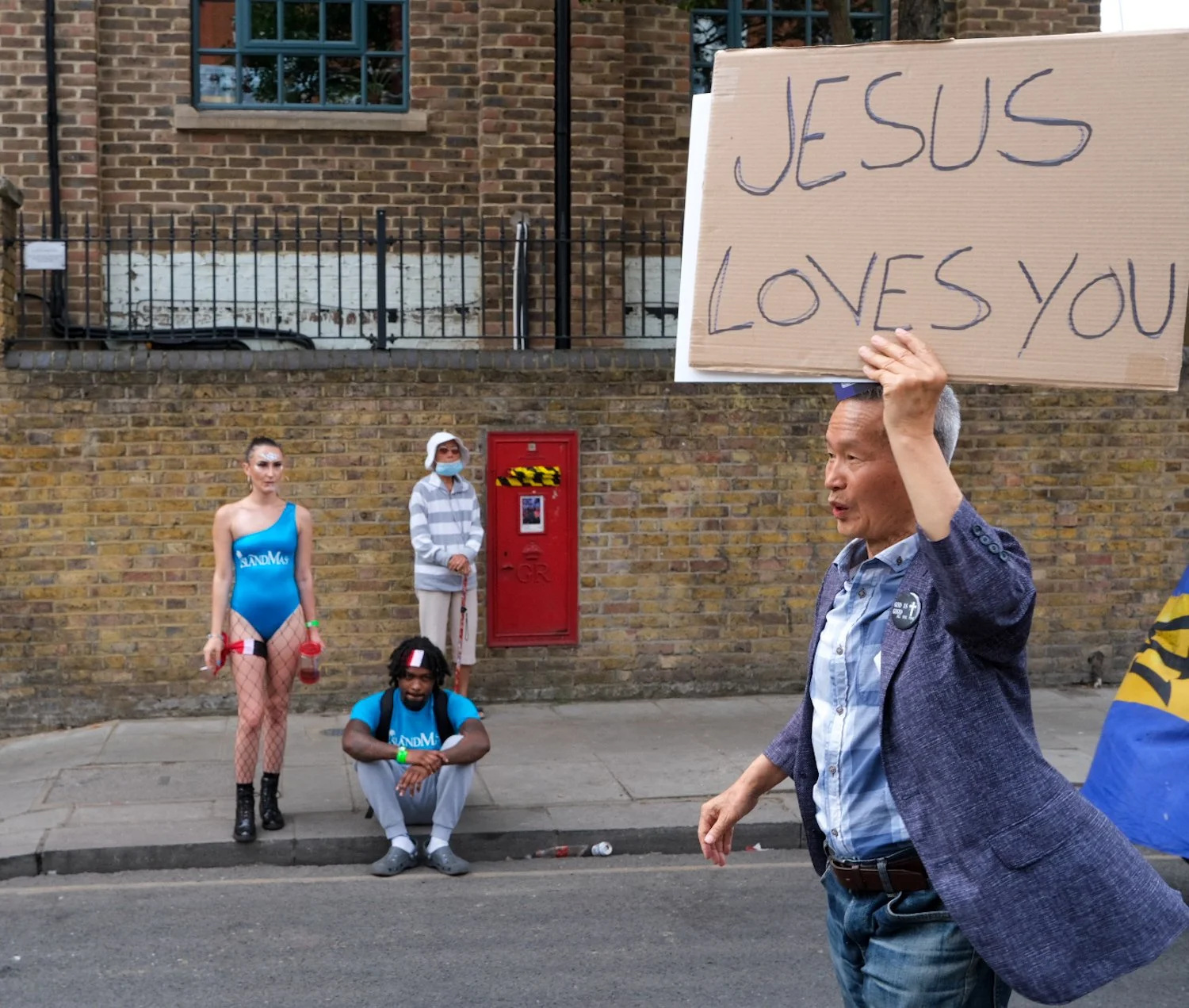 A man holding a large cardboard sign that reads 'JESUS LOVES YOU' in a street scene. Behind him, a woman in a blue swimsuit, fishnet stockings, and with makeup, stands next to a seated man in a blue shirt with 'SandM' written on it, and an older woma