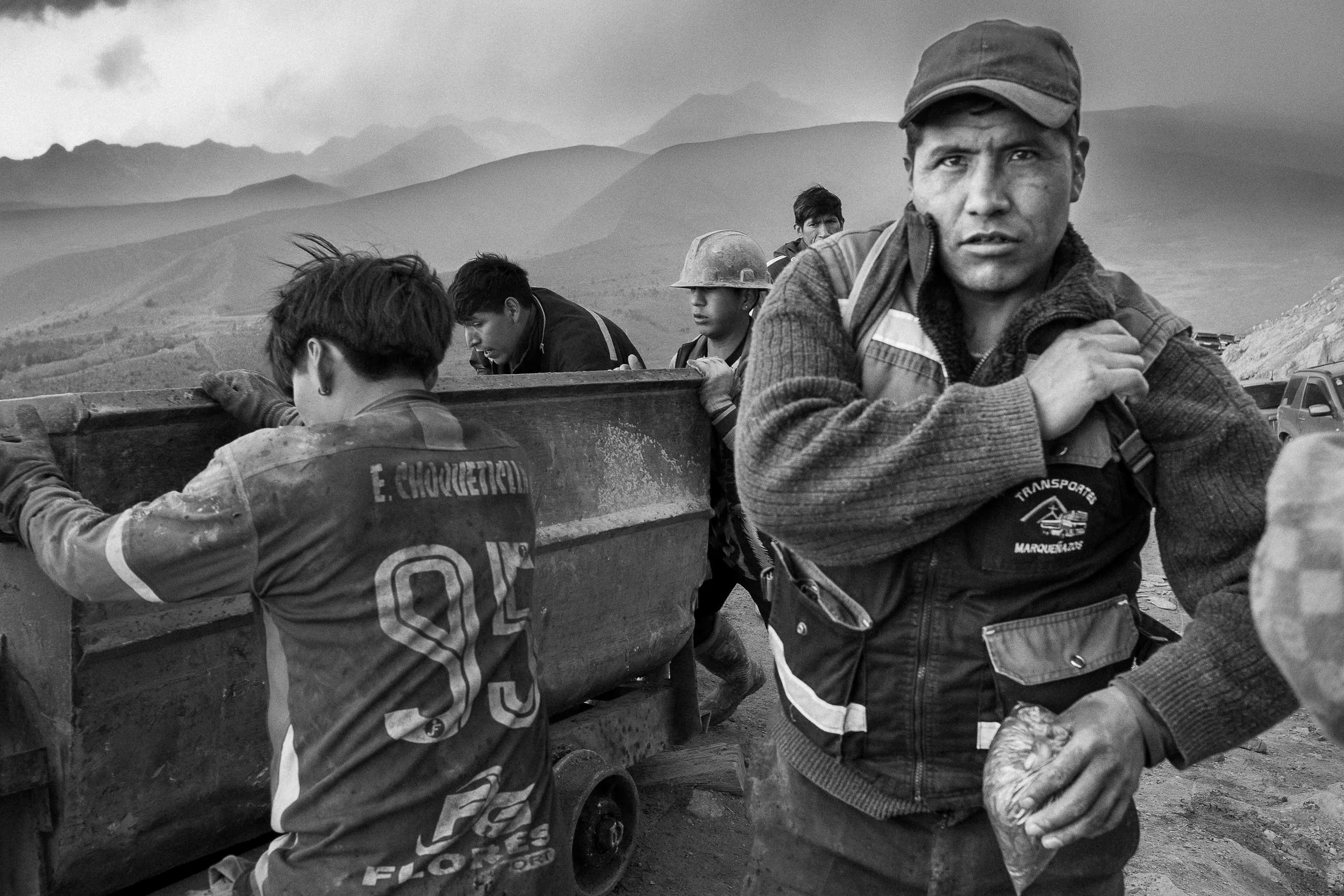 Black and white photo of several workers, some wearing helmets, with a mountainous landscape in the background, loading or unloading goods.
