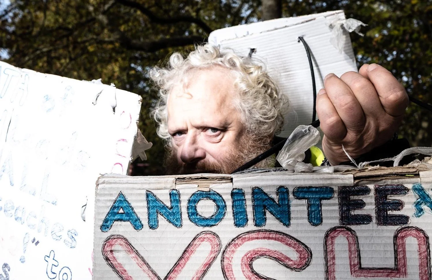 A man with curly white hair and a beard holding a protest sign, with only his face visible behind the sign. The sign says 'A NONE E' with additional text partially obscured.