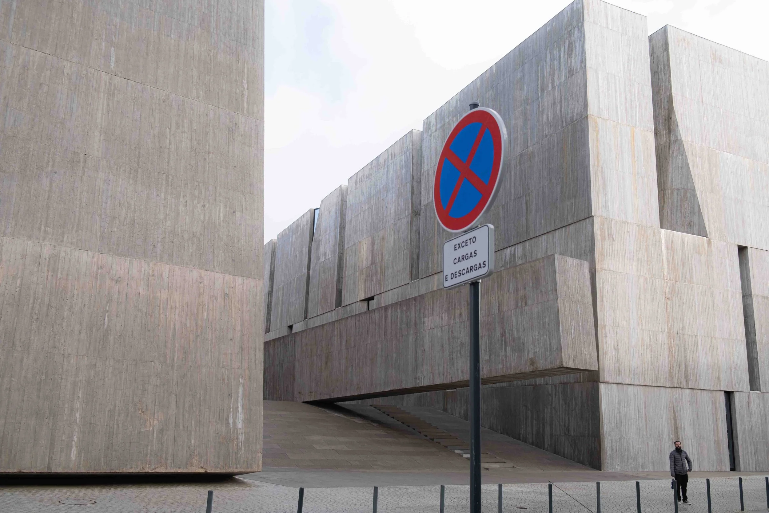 A modern building with large beige concrete blocks and a no-parking sign in the foreground, with a person standing in the lower right corner.