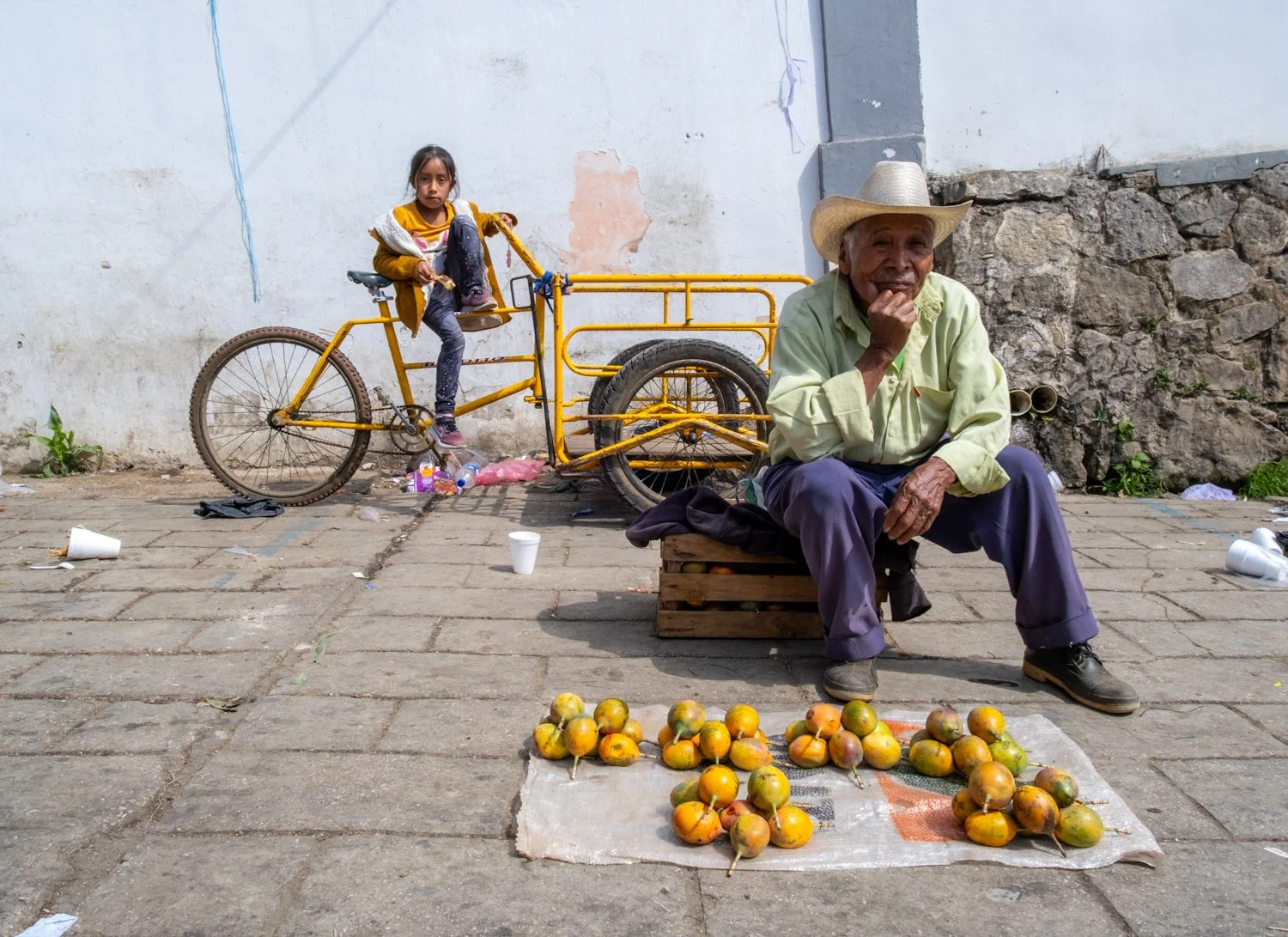 A young girl sitting on a yellow bicycle and an older man sitting on a wooden crate, selling oranges on the sidewalk with a wall and stone background.