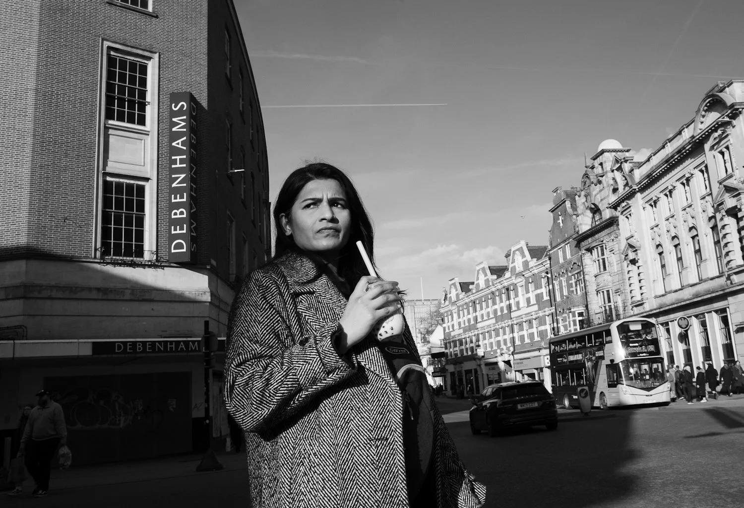 A woman with dark hair and a patterned coat standing on a city street holding a drink with a straw, with buildings, a double-decker bus, and pedestrians in the background.