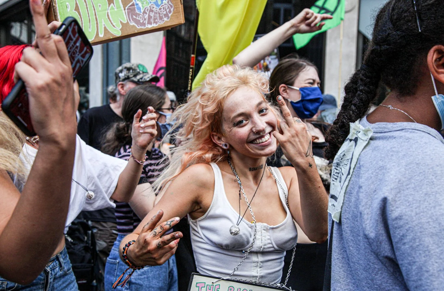 A smiling woman with blonde curly hair making a peace sign at a crowded outdoor event. She has numerous tattoos, piercings, and is wearing a white top with jewelry. Other people around her are wearing masks and appear to be enjoying the event.
