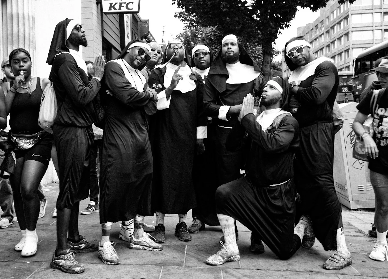 A group of people dressed in religious nun costumes, standing on a city street, some with sunglasses, and one person kneeling with hands together in prayer pose. Other pedestrians are visible in the background.