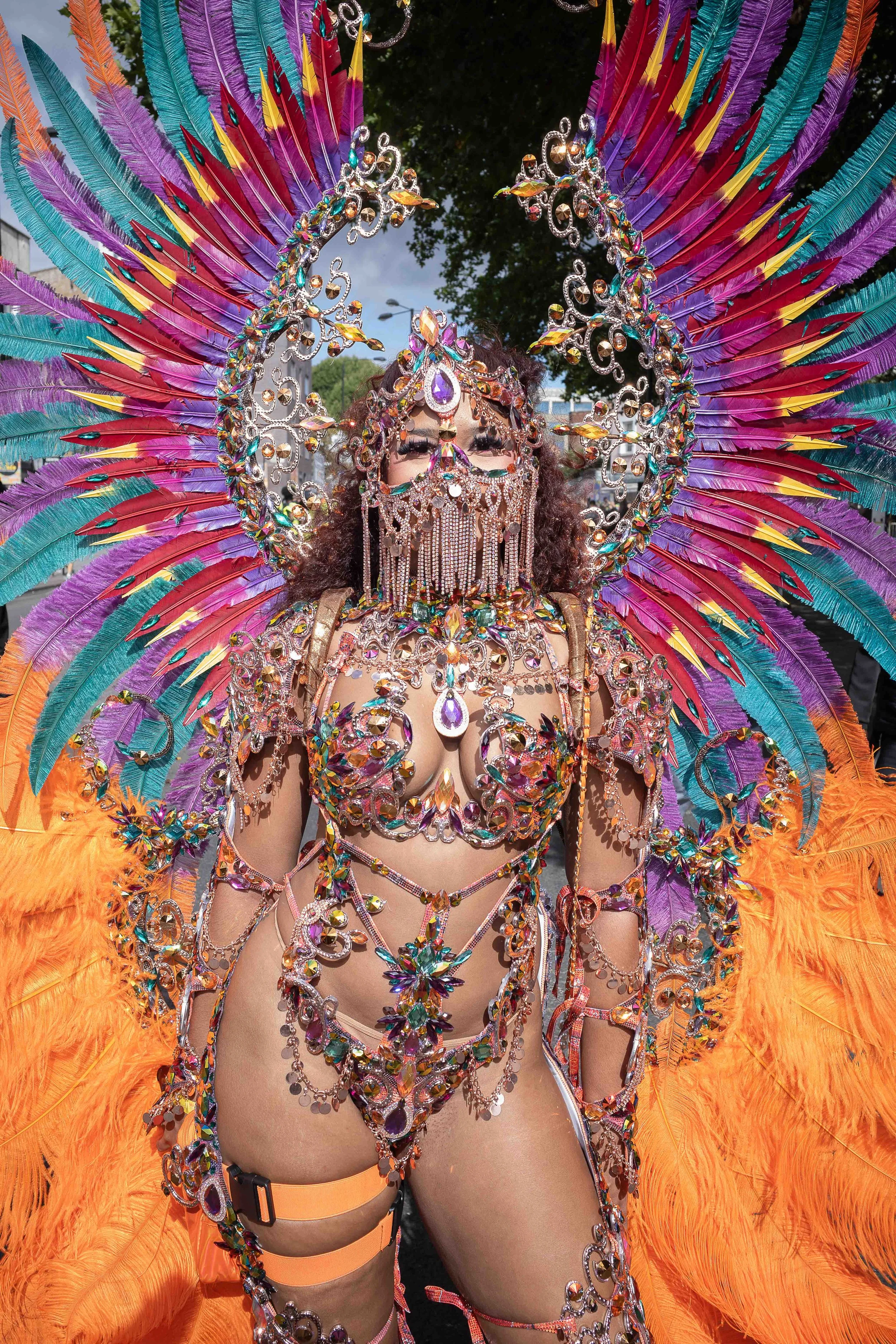 Person dressed in a colorful, elaborate costume with feathers, rhinestones, and sequins, wearing a mask with a bejeweled face covering, during a parade or festival.