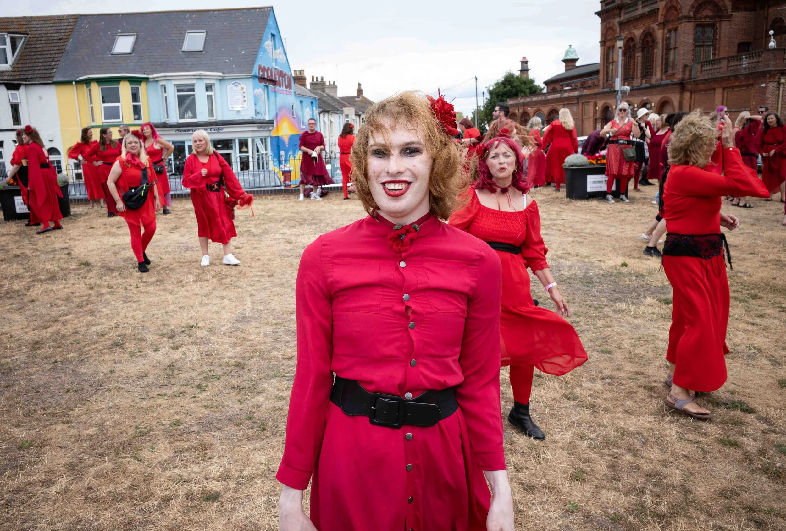 Group of people dressed in red clothing, standing and interacting in an outdoor area with grass and buildings in the background.