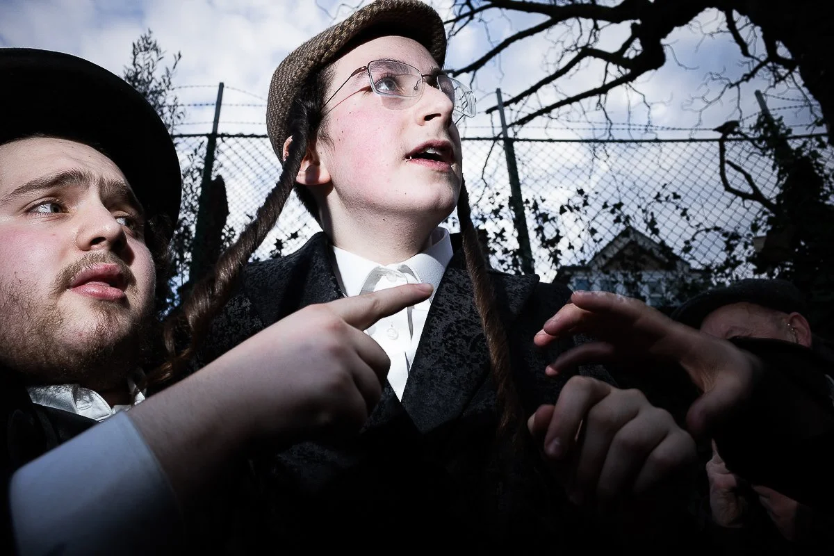 Three children dressed in costumes, one with glasses and a hat, outdoors near a chain-link fence and leafless trees.