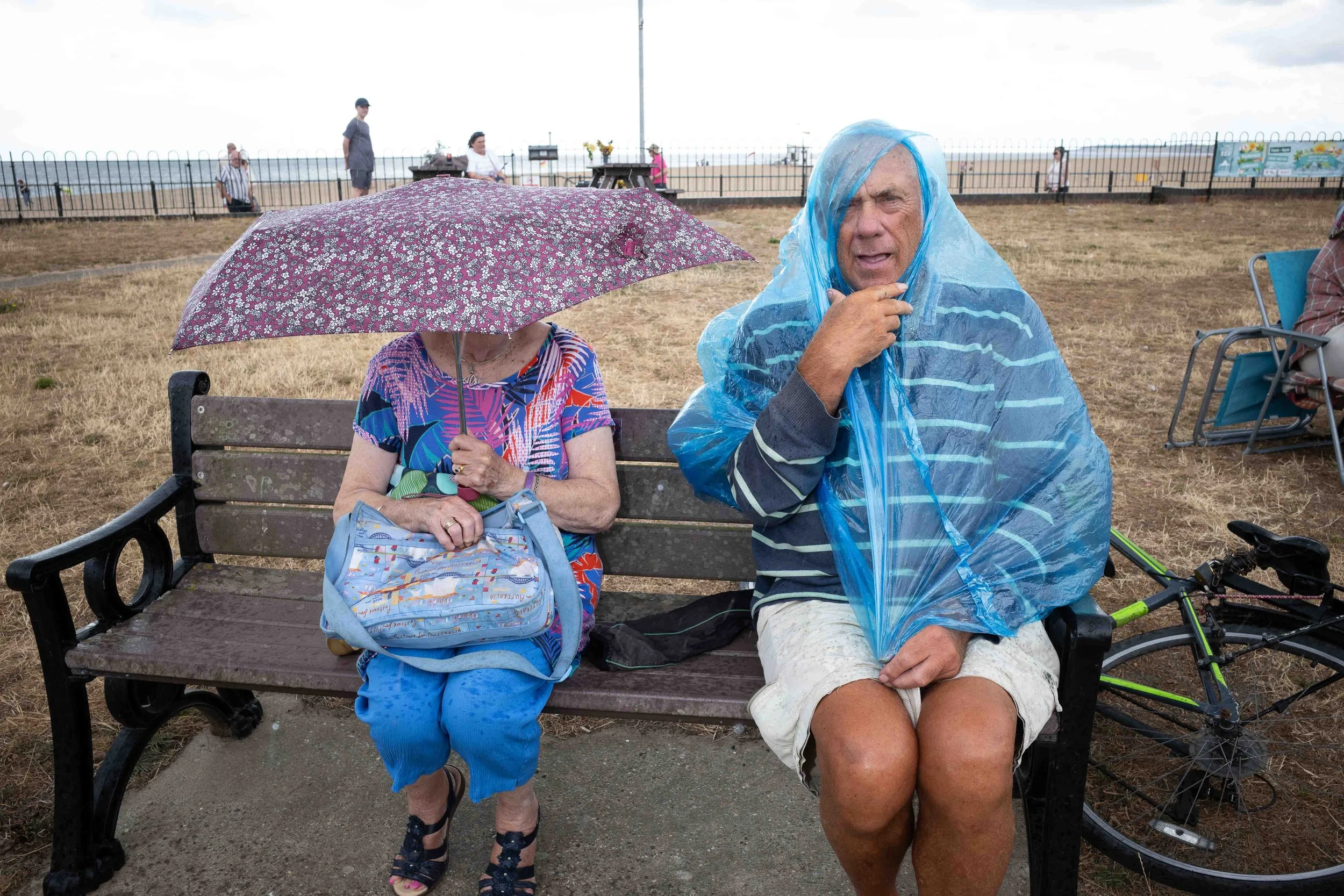 An elderly woman and man sit on a park bench, both wearing rain protection; the woman holds a floral umbrella, and the man wears a blue rain poncho. They are at an outdoor event near the beach, with a fenced area, people walking, and a table in the b