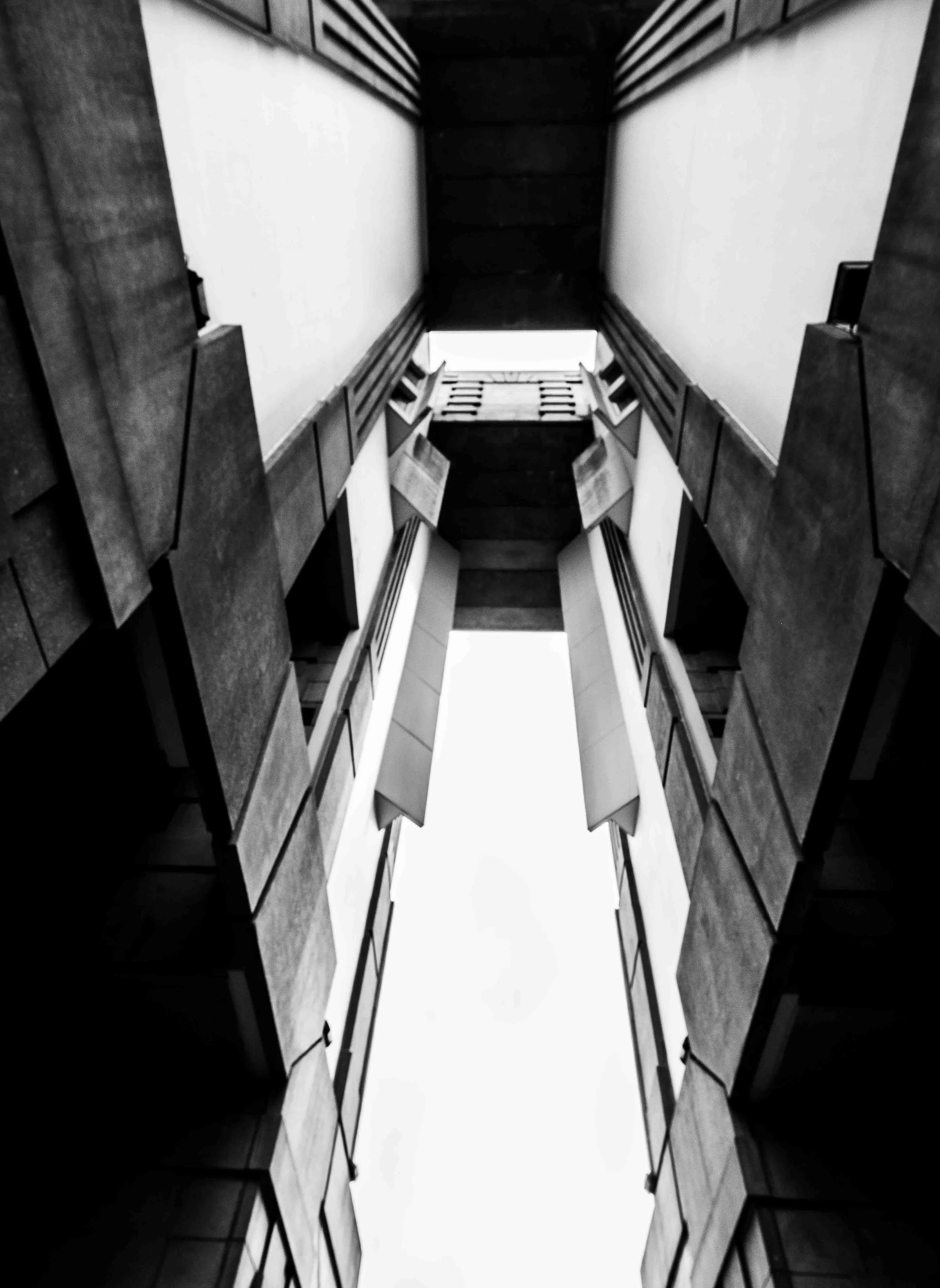 Black and white photo of a modern building with a narrow view looking up between two parallel structures with a gap of sky in the center.