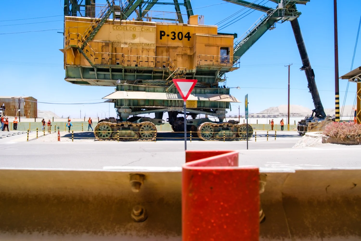 A large yellow mining or construction machine on a road, with a crane arm attached, surrounded by construction workers and safety barriers, in a desert environment with mountains in the background.