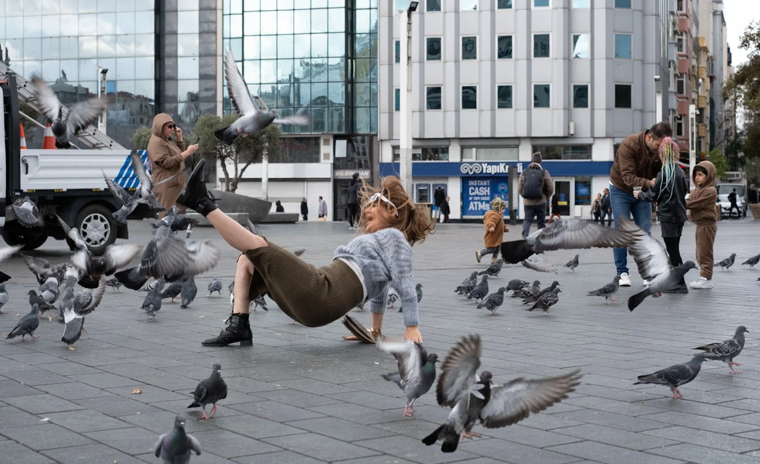A woman lying on her back on a city sidewalk, surrounded by pigeons, with some birds flying around her. In the background, a man and two children are interacting, and other people are walking on the street.