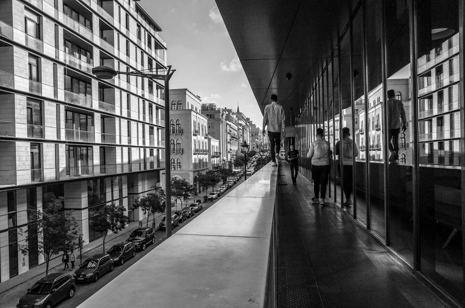 A black and white photo shows a city street scene with tall buildings, cars parked along the curb, and pedestrians walking on a balcony or walkway. The focus is on the reflection of people walking in glass windows of a building on the right.