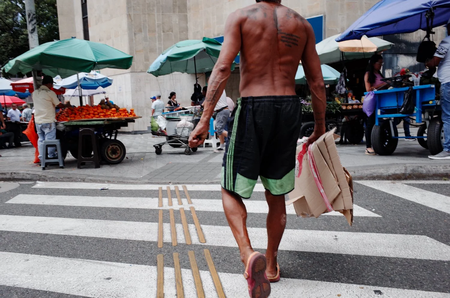 A shirtless man with tattoos, wearing black and green shorts, walking across a street holding a shopping bag and cardboard boxes in a busy market with vendors and umbrellas.