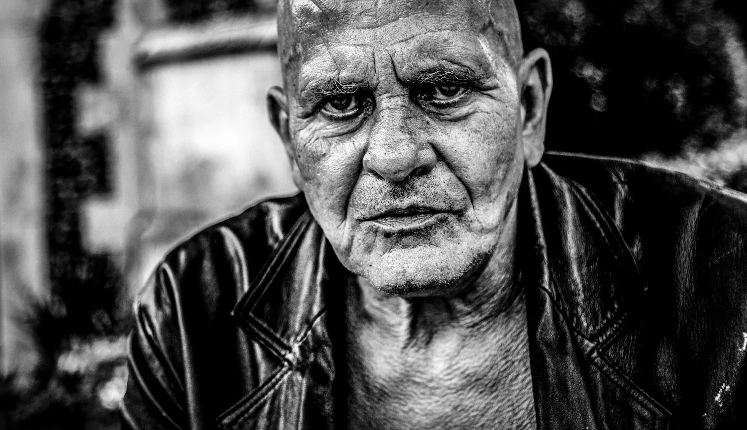 Close-up black and white photograph of an elderly man with a rugged face, intense expression, and wearing a leather jacket.