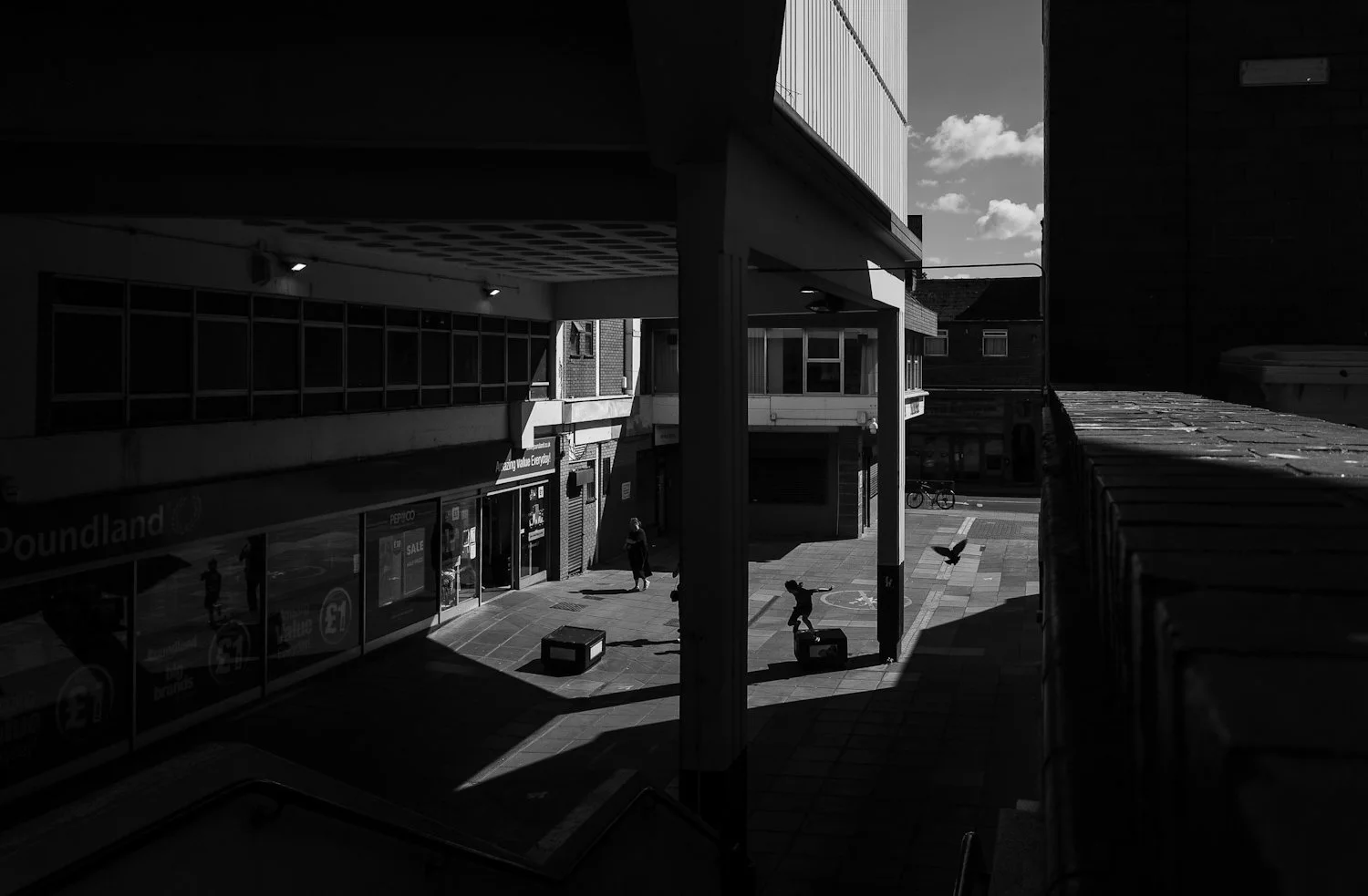 Black and white photo of a city street scene with people walking, a bicycle, and a pigeons, with buildings and a storefront named 'Soundland' visible.