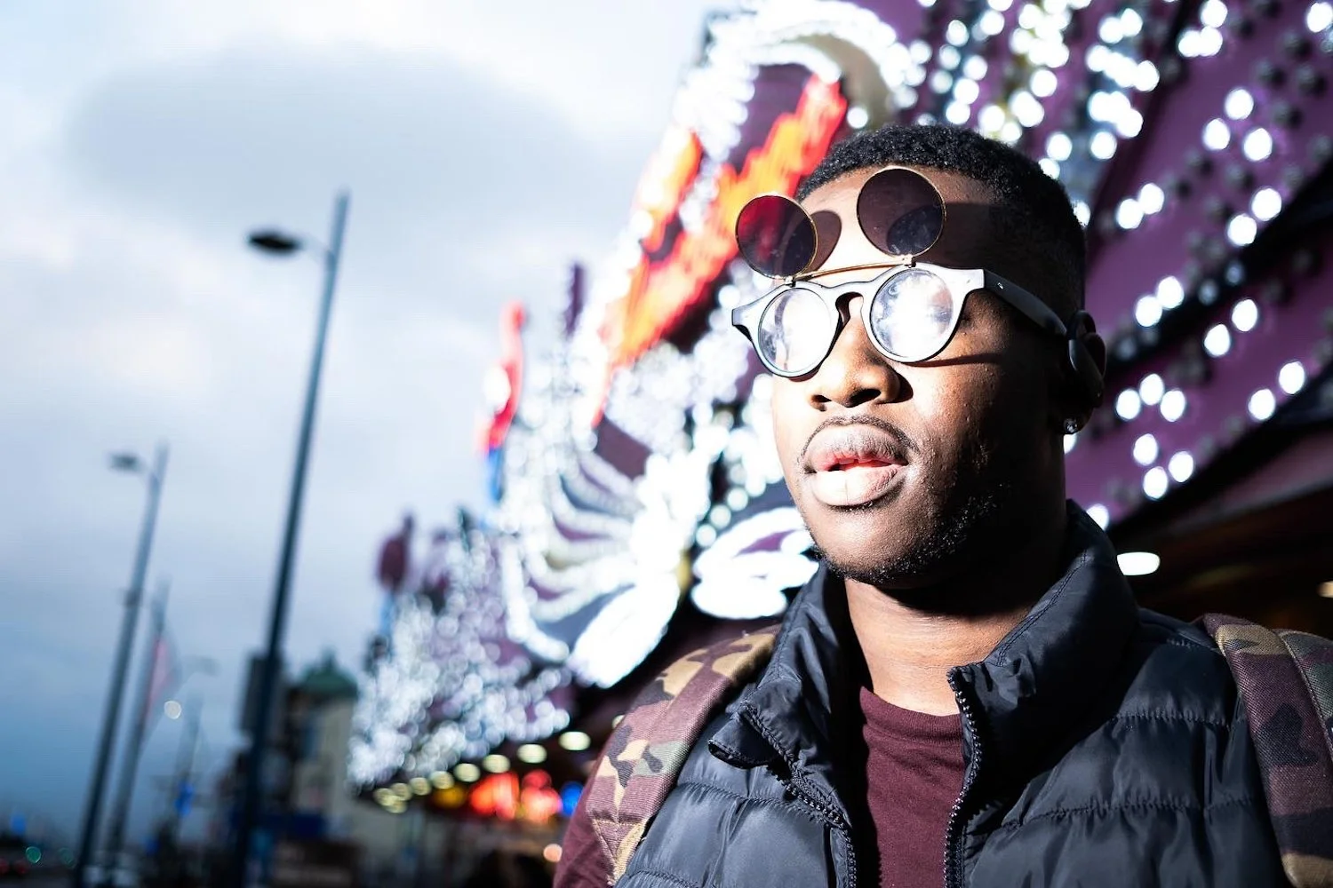A man wearing sunglasses with multiple lenses photoshopped over his face, standing outdoors in front of a brightly lit carnival or amusement park ride, with a cloudy sky overhead.