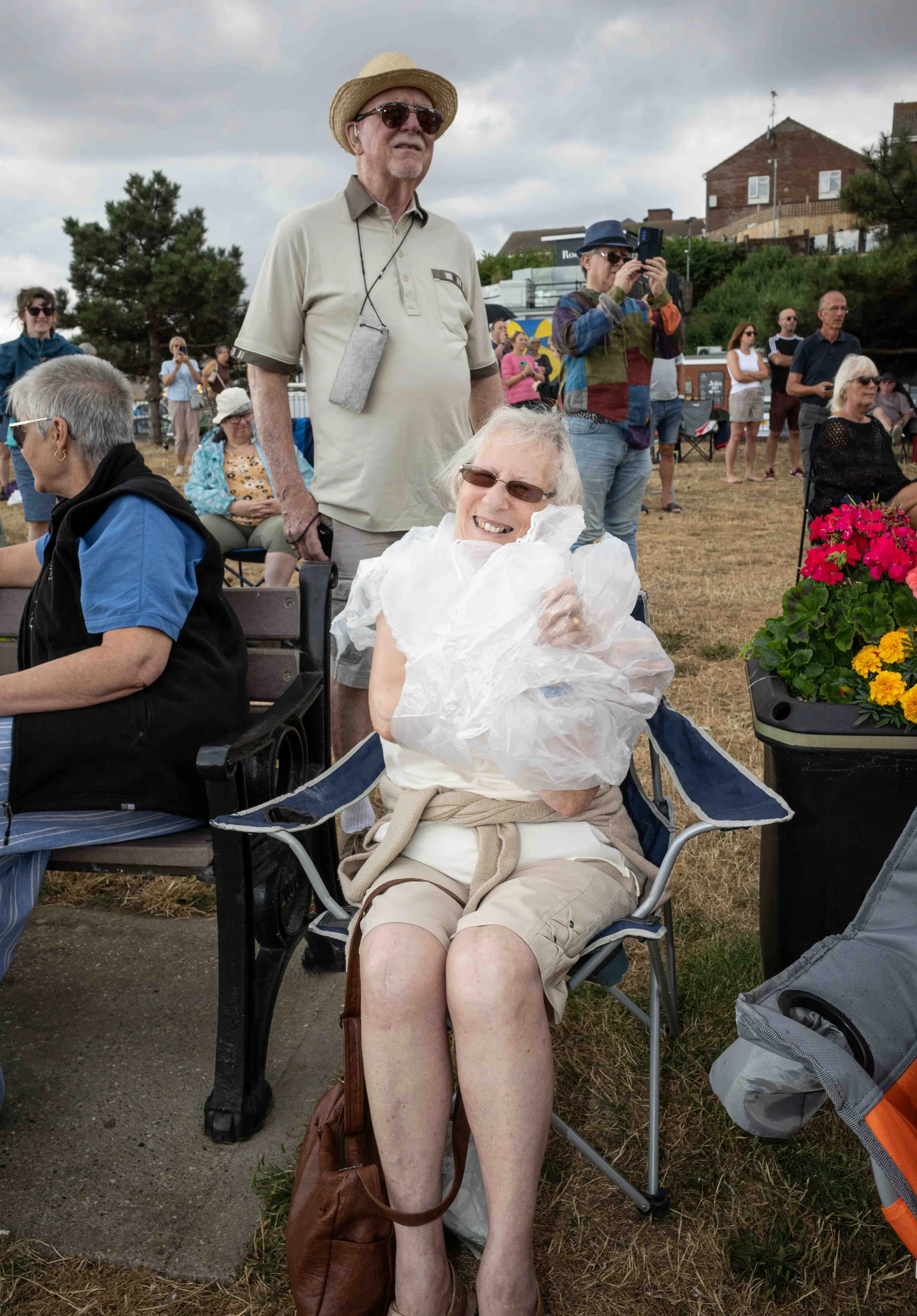 An elderly woman with short gray hair wearing sunglasses and a white blouse seated in a folding chair, smiling. A brown purse rests at her feet and a brown bag is hanging from her chair. A group of people are standing and sitting outdoors on a grassy