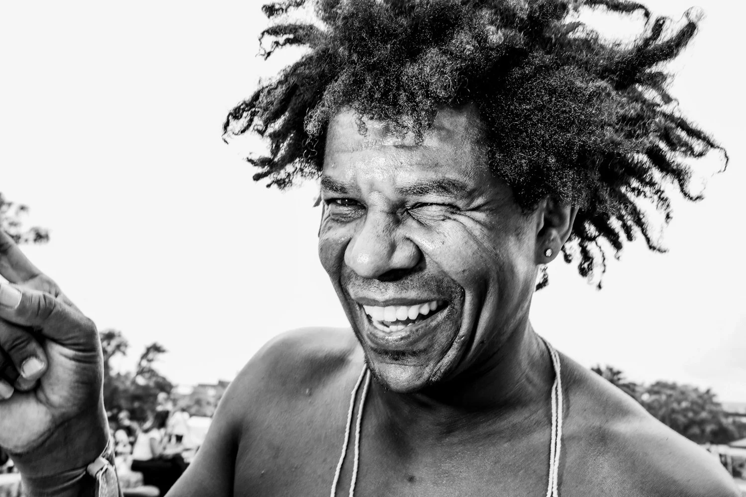 A man with curly hair winks and smiles widely, showing his teeth, while holding his hand up. He is shirtless and has an earring in his left ear, wearing necklaces, with a background of blurred people and trees.