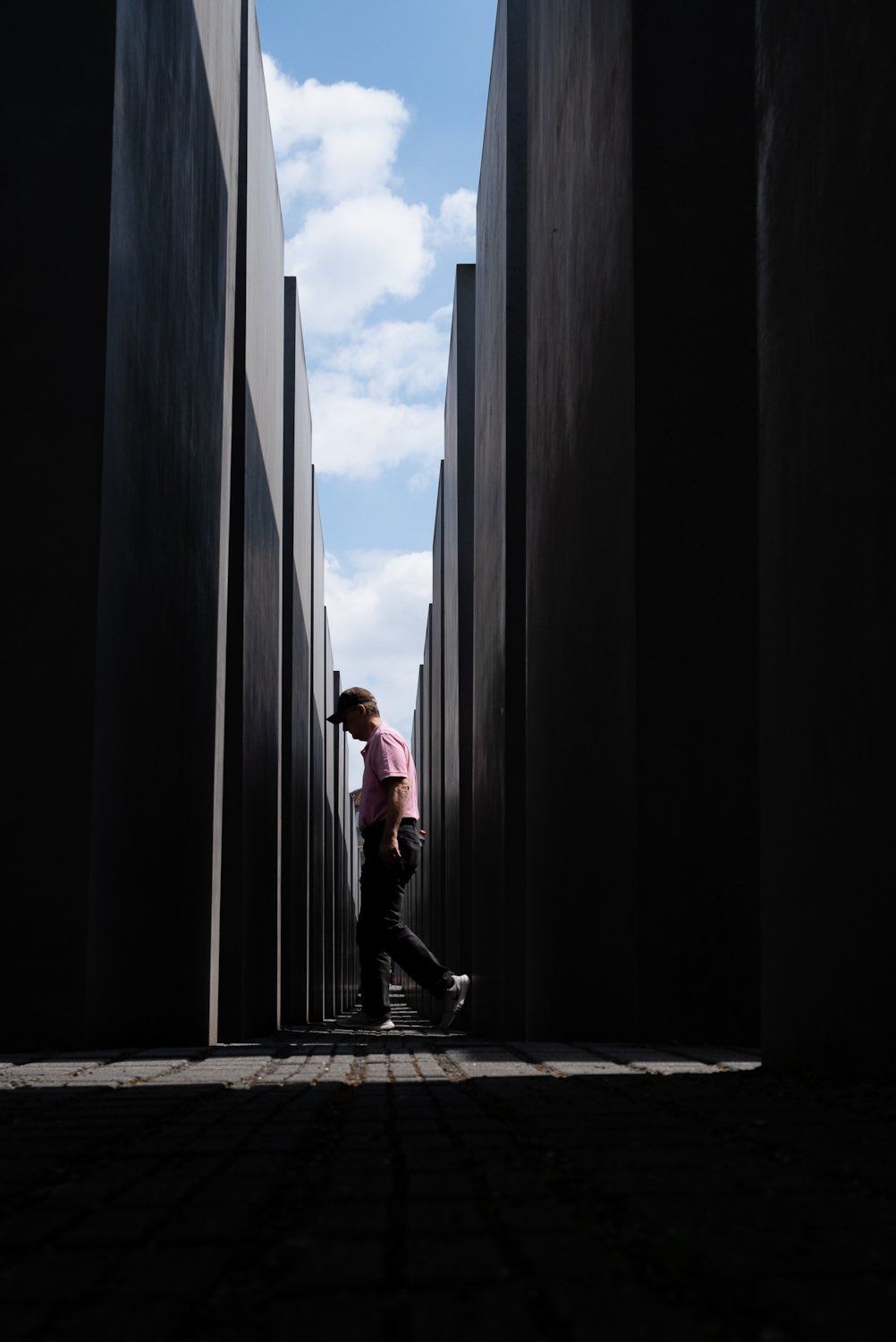 A person wearing a pink shirt, black pants, and a cap walking through the Holocaust Memorial in Berlin, with tall concrete concrete slabs on either side and a partly cloudy sky above.
