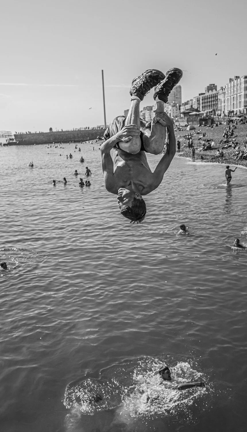 A person is performing a backflip into the water at a crowded beach, with many people swimming and lounging on the shore, and buildings in the background.