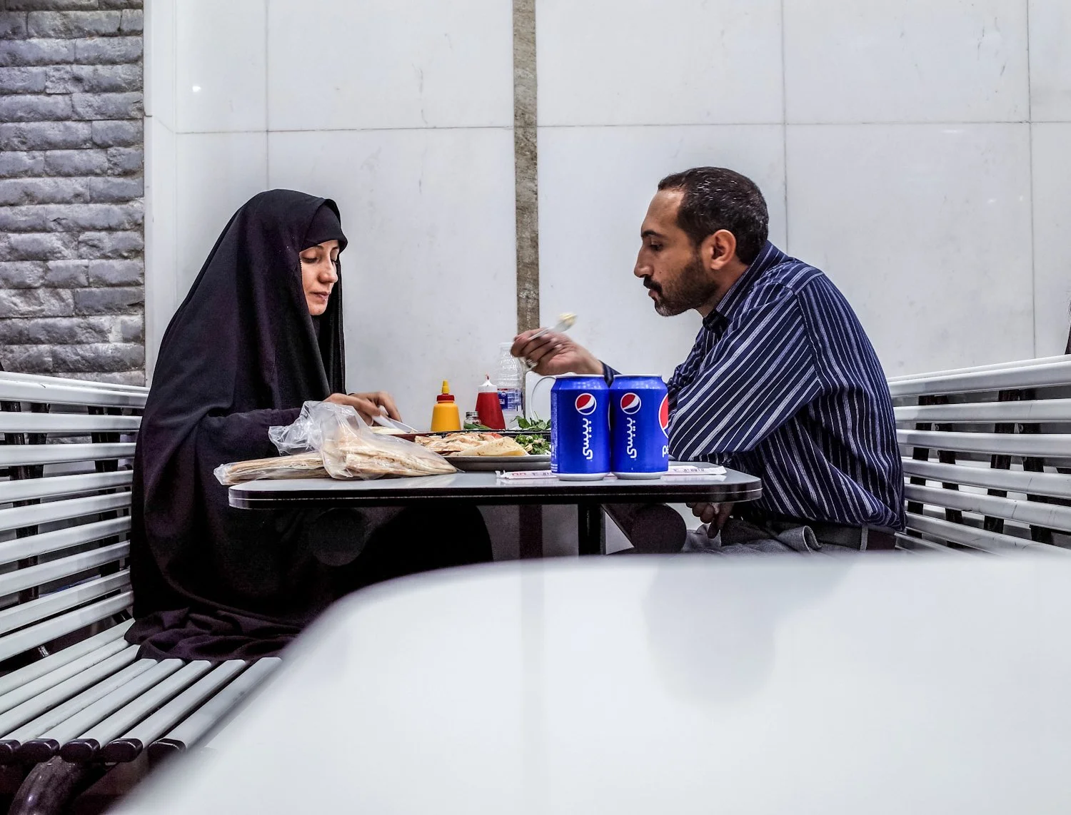 A man and a woman sit at a dine-in restaurant table, having a meal. The woman wears a black hijab and dark clothing, while the man wears a striped shirt. The table has two cans of Pepsi, condiments, a pizza, and a plastic bag with food.