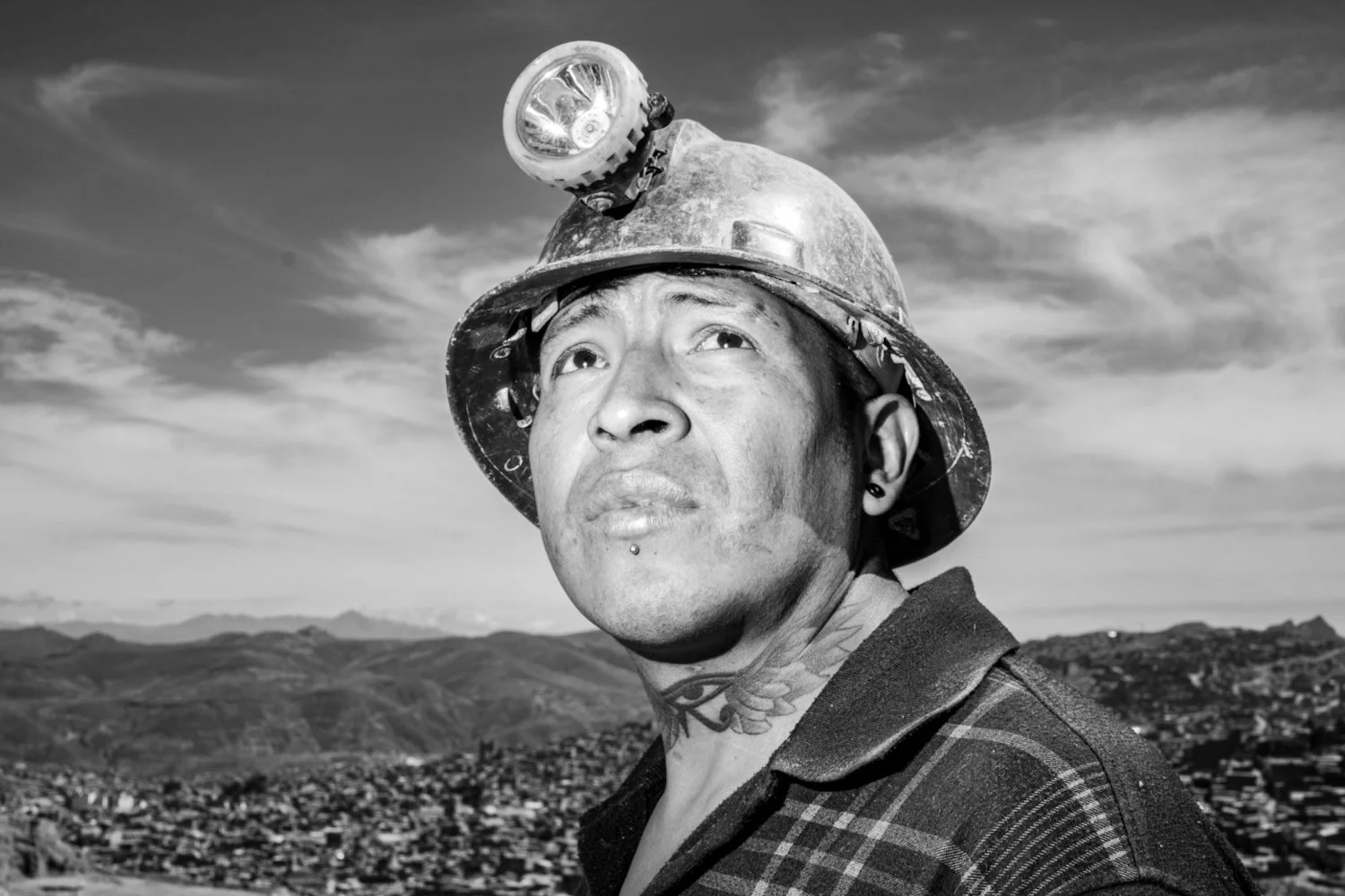 Black and white photo of a male miner wearing a helmet with a headlamp, looking up with a thoughtful expression, with a mountainous landscape in the background.
