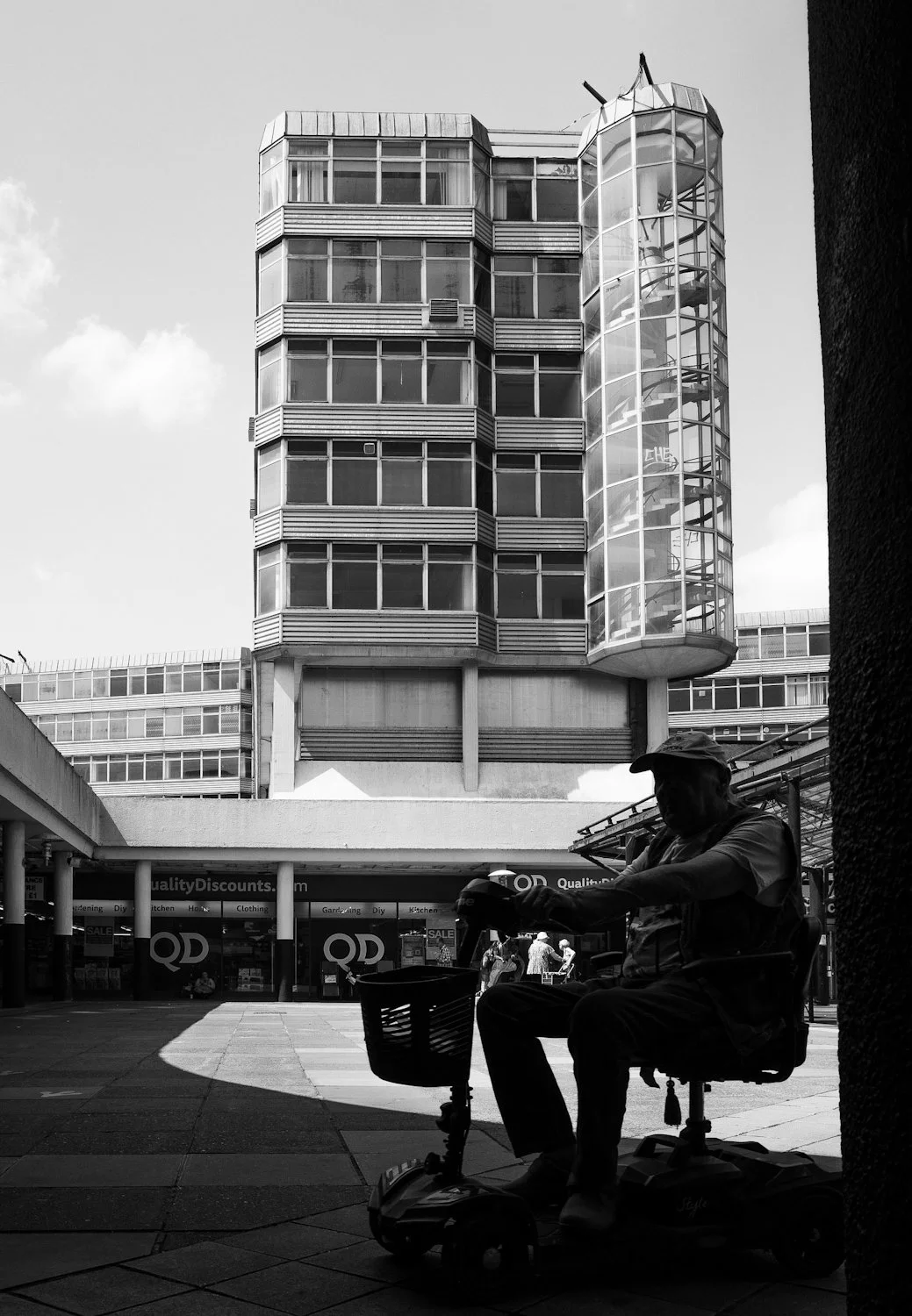 A silhouette of a man in a cap sitting on an electric scooter with a shopping basket, with a modern multi-story building with glass windows and a circular staircase in the background.