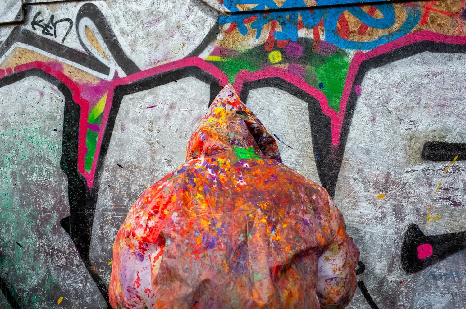A person wearing a hooded jacket covered in multicolored paint stands in front of a graffiti-covered wall, with their back to the camera.