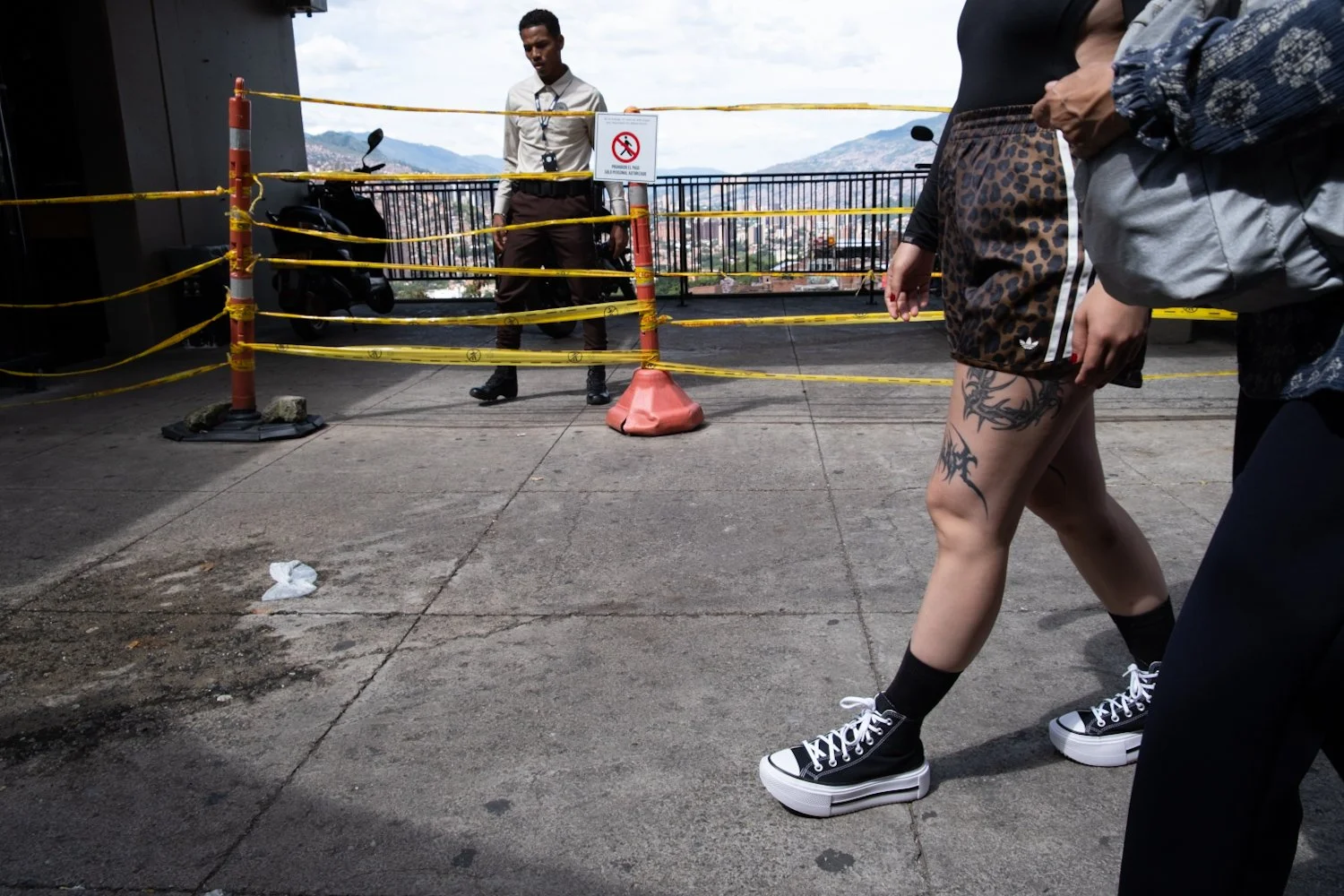 People walk past a yellow caution tape blocking an area on a rooftop with a city view in the background. A person with tattoos wearing platform shoes and a skirt is in the foreground.