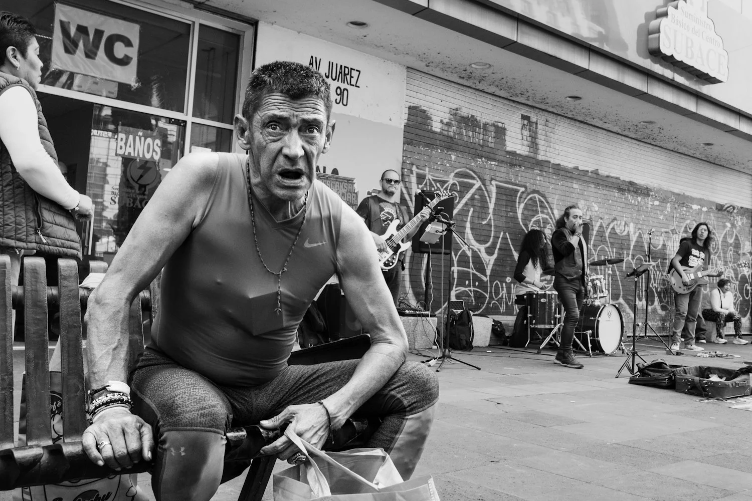 A man in athletic wear leaning forward on a bench near a street band, with a music band performing in the background on a city sidewalk, including a guitarist, drummer, and a vocalist.