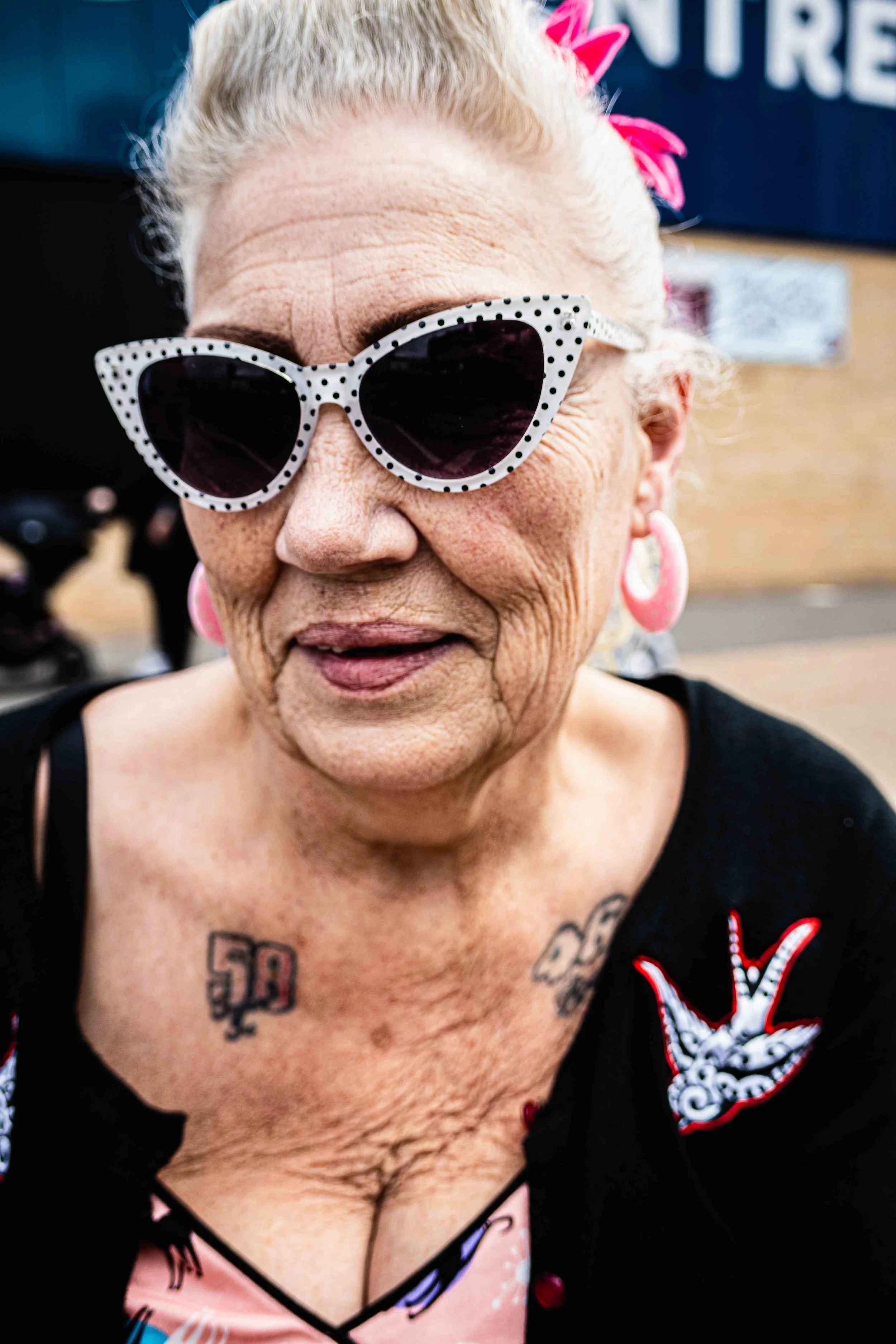 Close-up of an elderly woman with white hair wearing white sunglasses with black polka dots and pink earrings. She has tattoos on her chest and is wearing a pink top and black outer garment with red buttons.