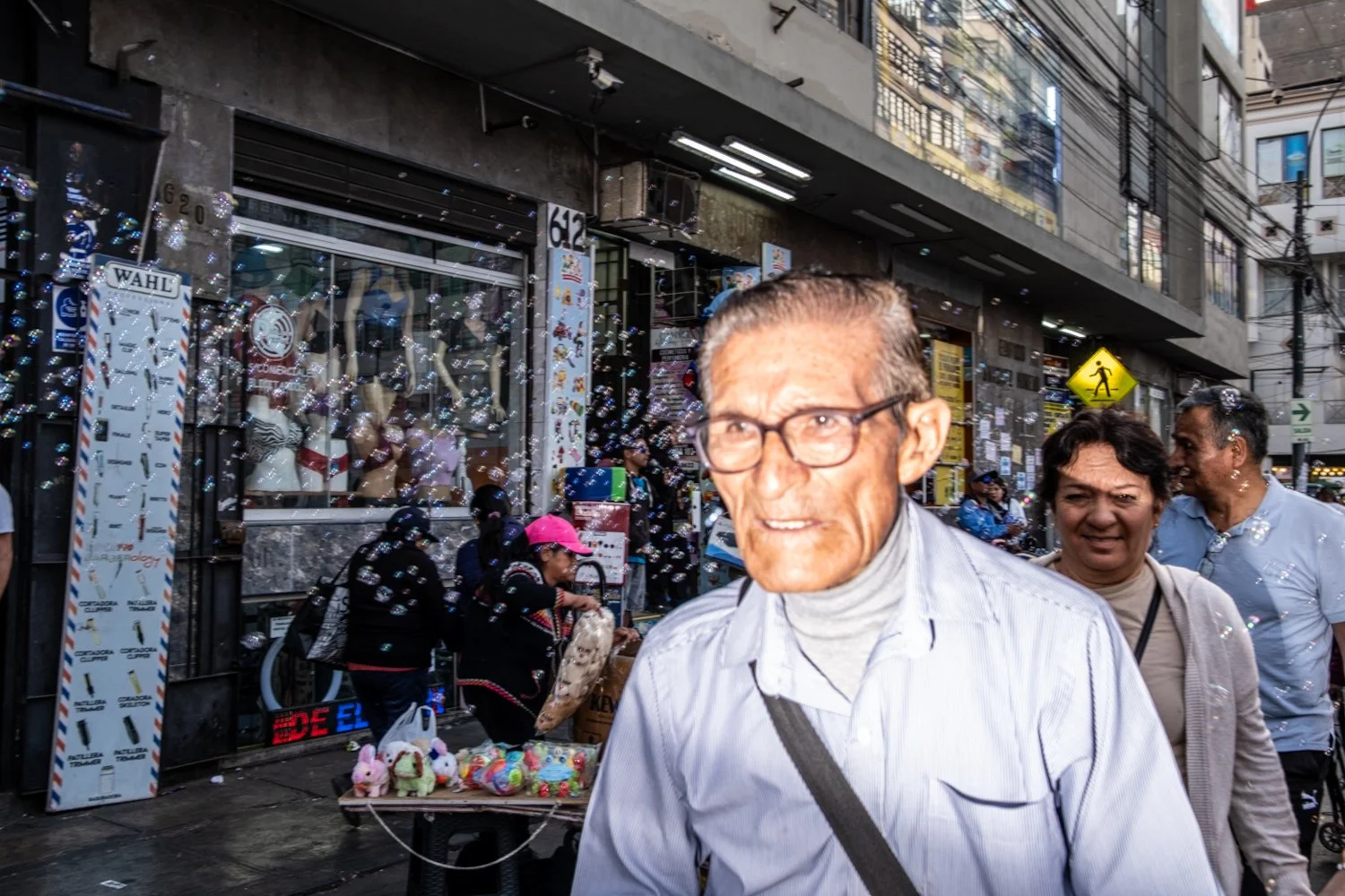 Street scene with a man wearing glasses and a white shirt in the foreground; other pedestrians and street vendors with stuffed toys on a table are visible in the background. There are bubbles in the air and retail storefronts behind them.