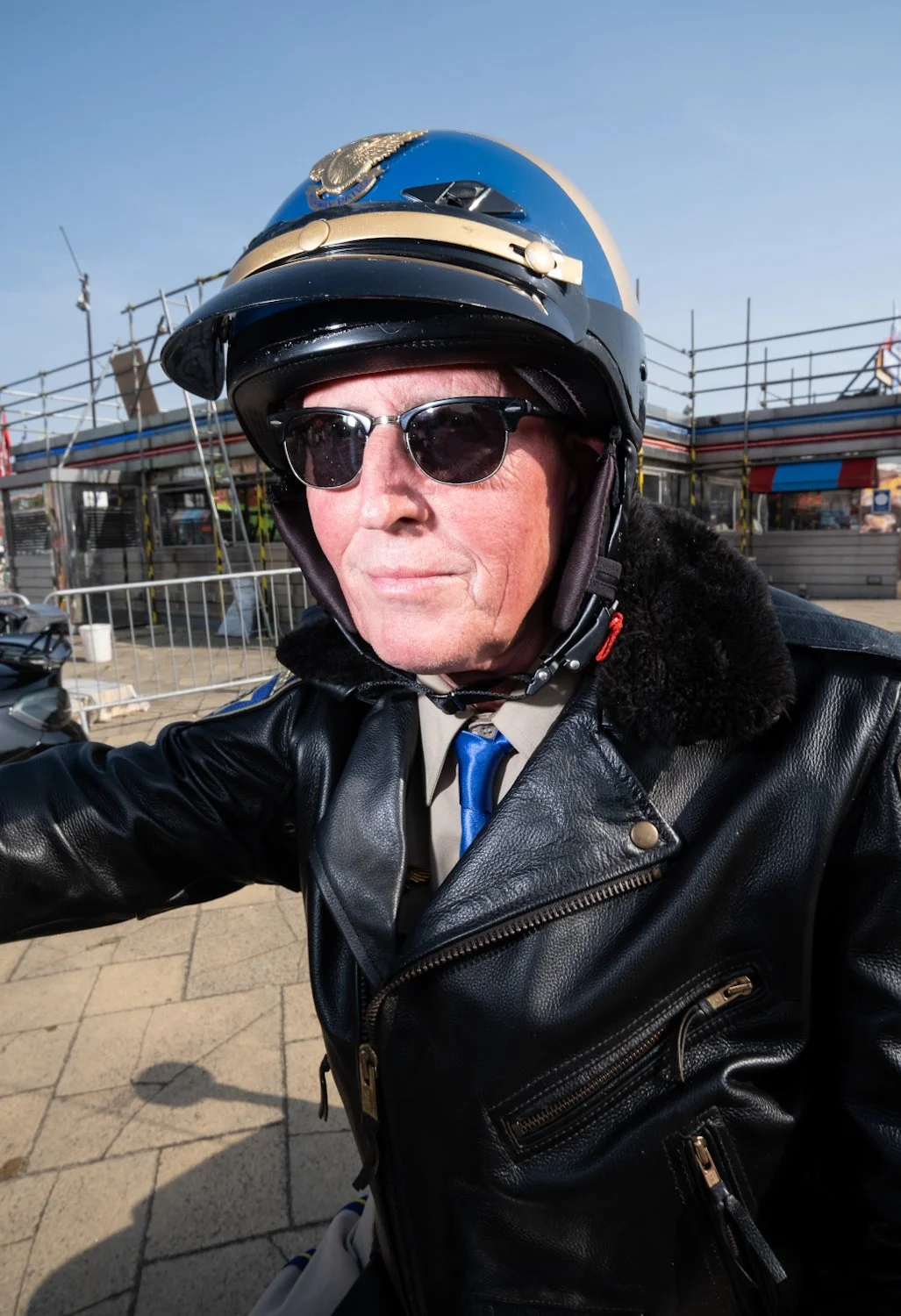 Man riding a motorcycle, wearing a black leather jacket, sunglasses, and a blue motorcycle helmet with a badge or emblem, at an outdoor location with buildings and fencing in the background.