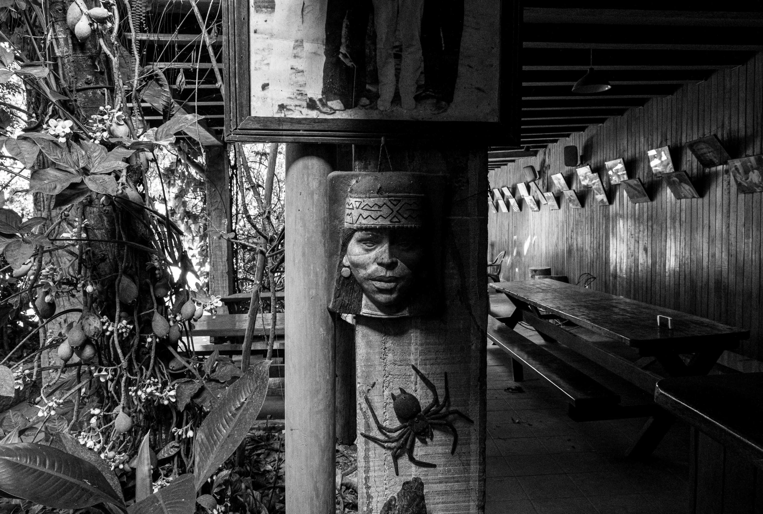 Black and white photo of a large carved wooden sculpture of a woman’s face and a hat, mounted on a wooden pillar. Behind it, there are plants with fruit, a mirror, a wooden wall decorated with framed photos, and a room with a wooden ceiling and shelv
