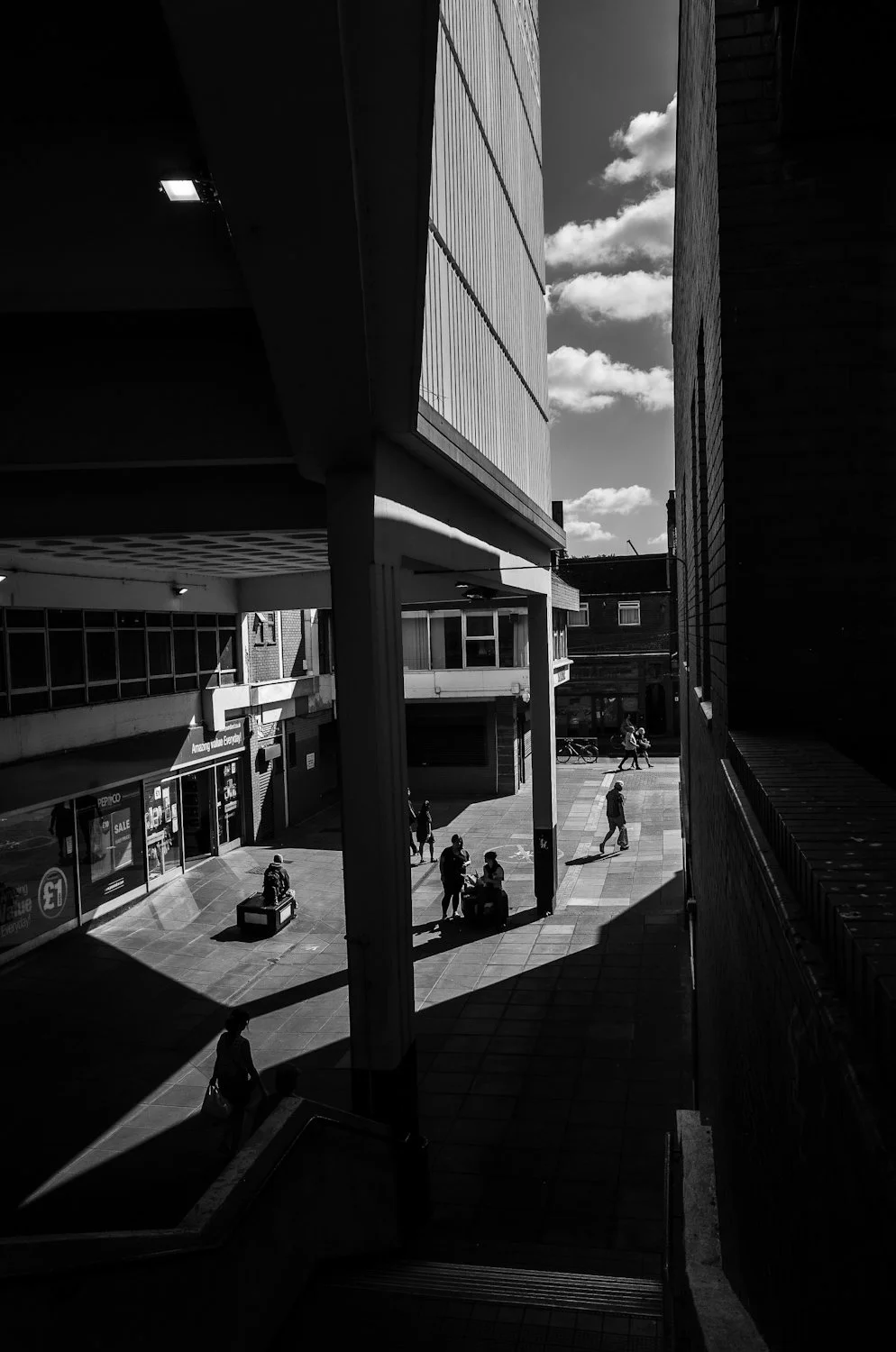 Black and white photo of a city street with people walking and sitting, buildings with large windows, and a partly cloudy sky.