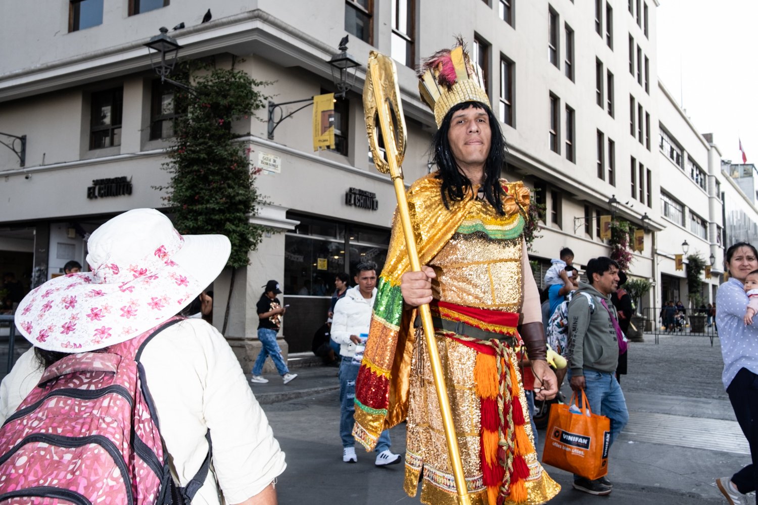 Person dressed as a regal figure in golden costume holding a staff, walking on a city street with onlookers around.