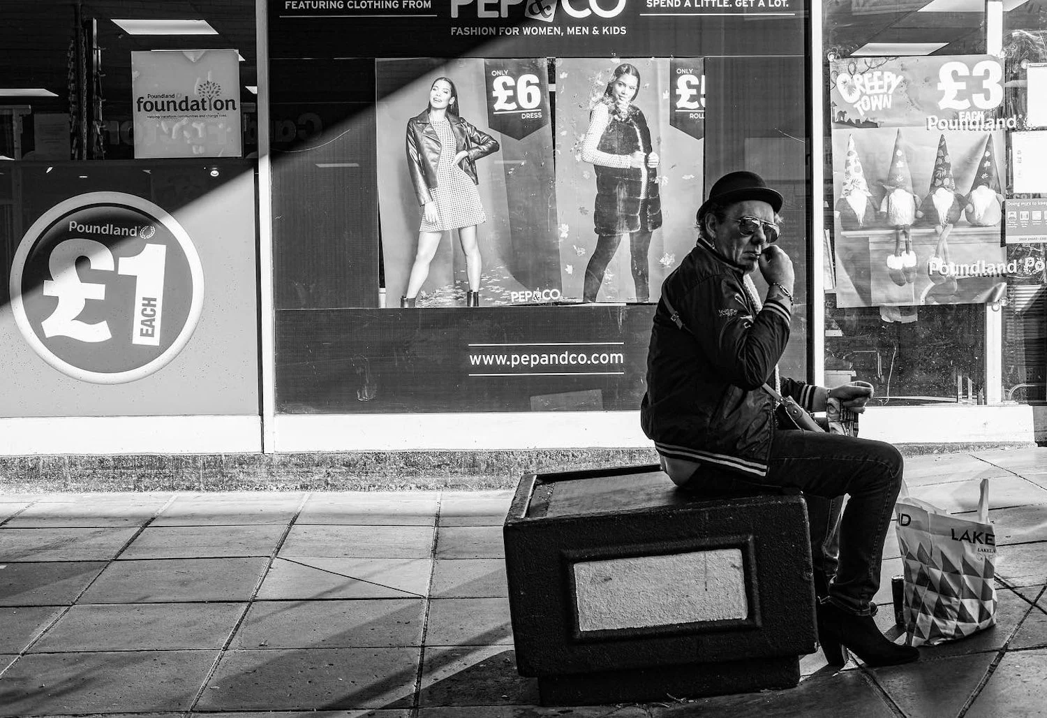 A man sitting on a bench outside a store, wearing sunglasses and a black jacket, appears to be smoking. The store window behind him displays posters of women in fashionable clothing. The man has a shopping bag on the ground beside him.