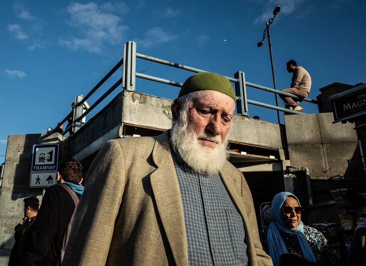 An older man with a white beard, wearing a green cap and a brown blazer, standing outdoors on a sunny day. There are a few other people, including a woman wearing sunglasses and a headscarf, behind him. In the background, there is an elevated walkway