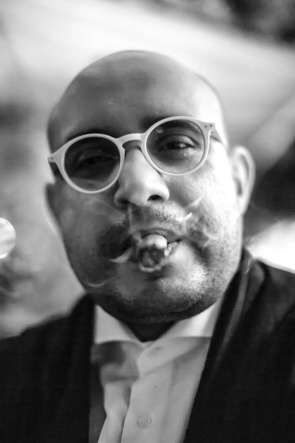 A black and white close-up of a man with a bald head and facial hair, wearing round sunglasses and a formal shirt, smiling and making a gesture with his hand.