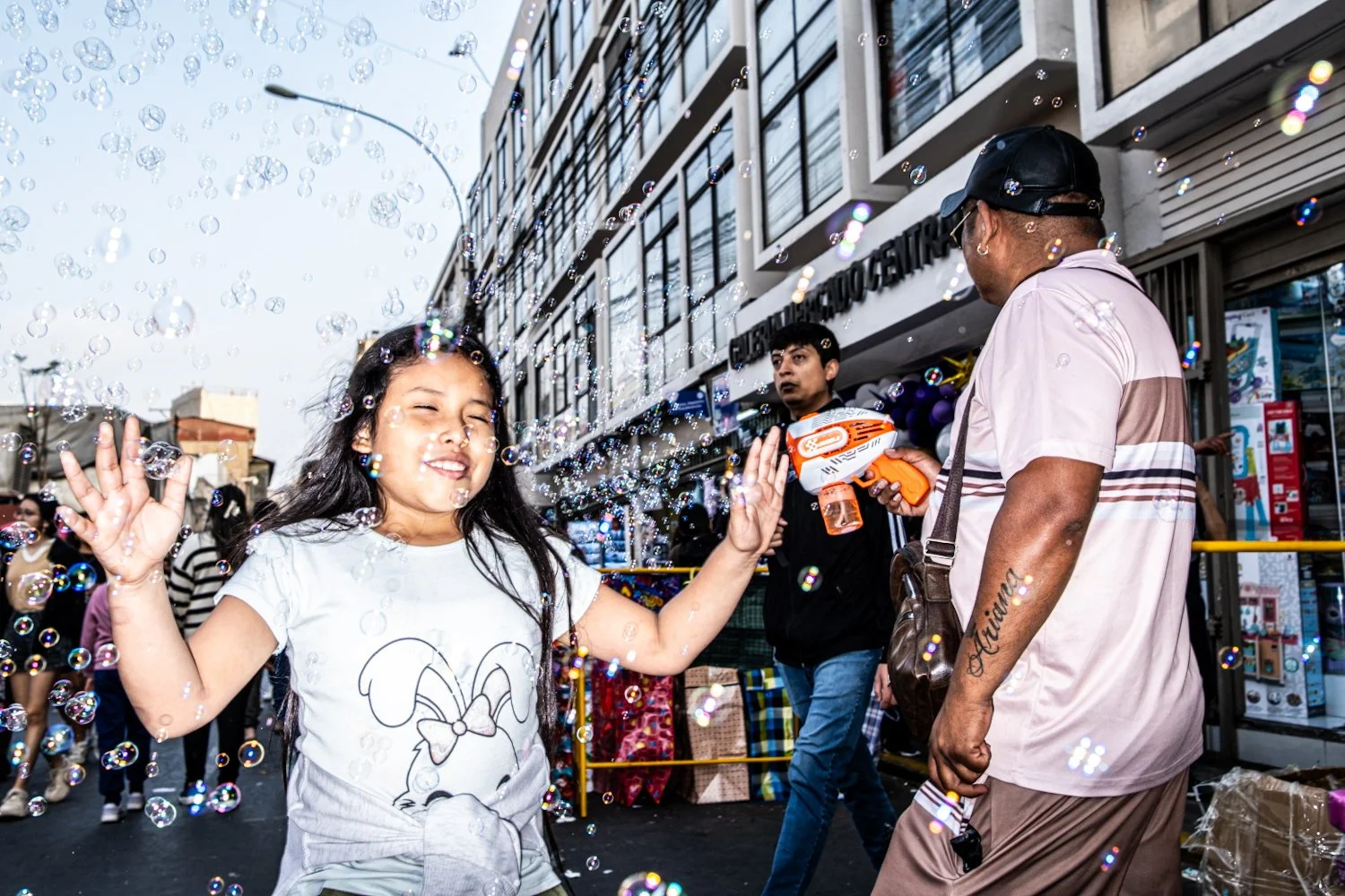 A young girl playing with bubbles on a city street, with a man holding a bubble gun and others in the background near a building.