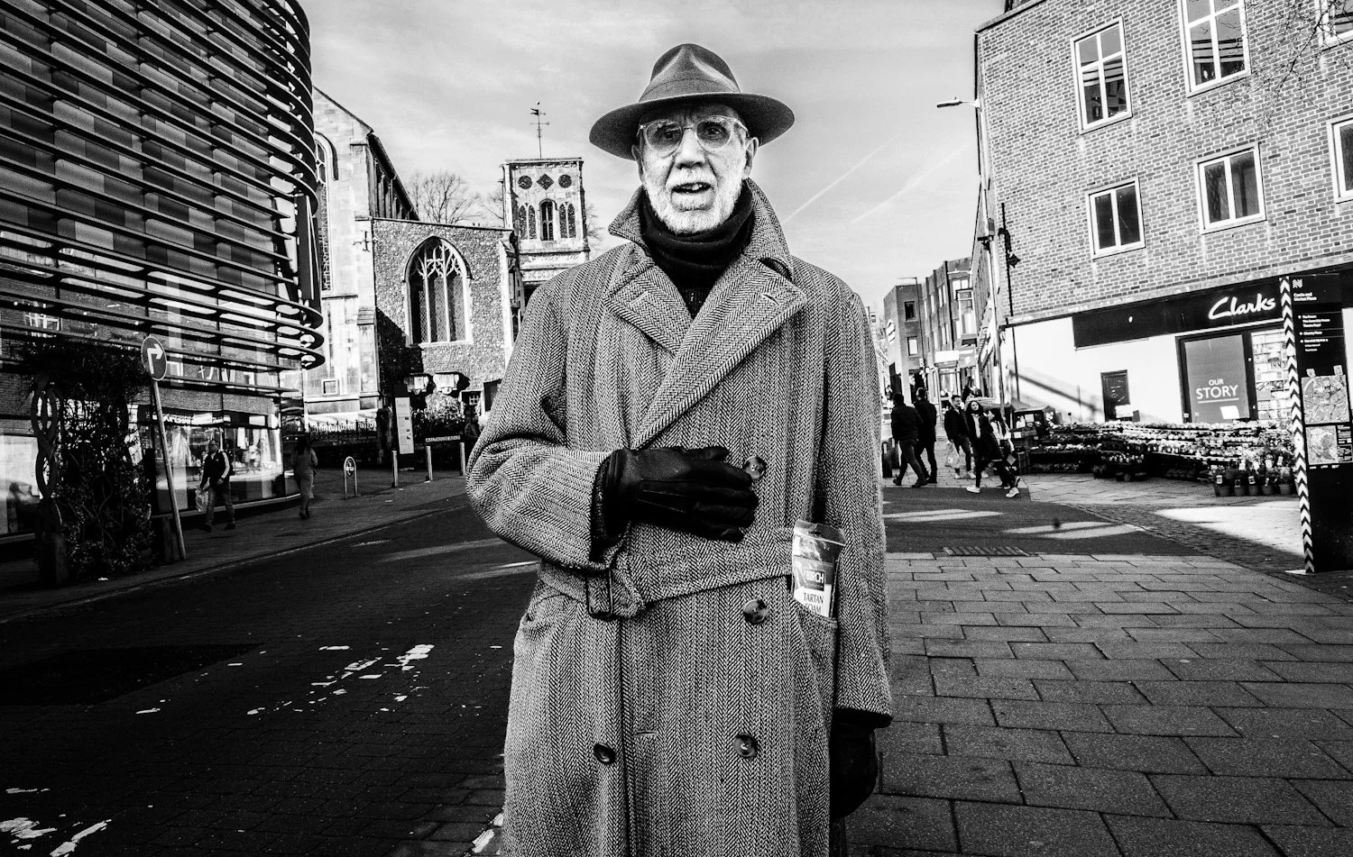 A man wearing a overcoat, gloves, and a hat is standing on a city street holding a cup and a small object, with several people walking in the background.