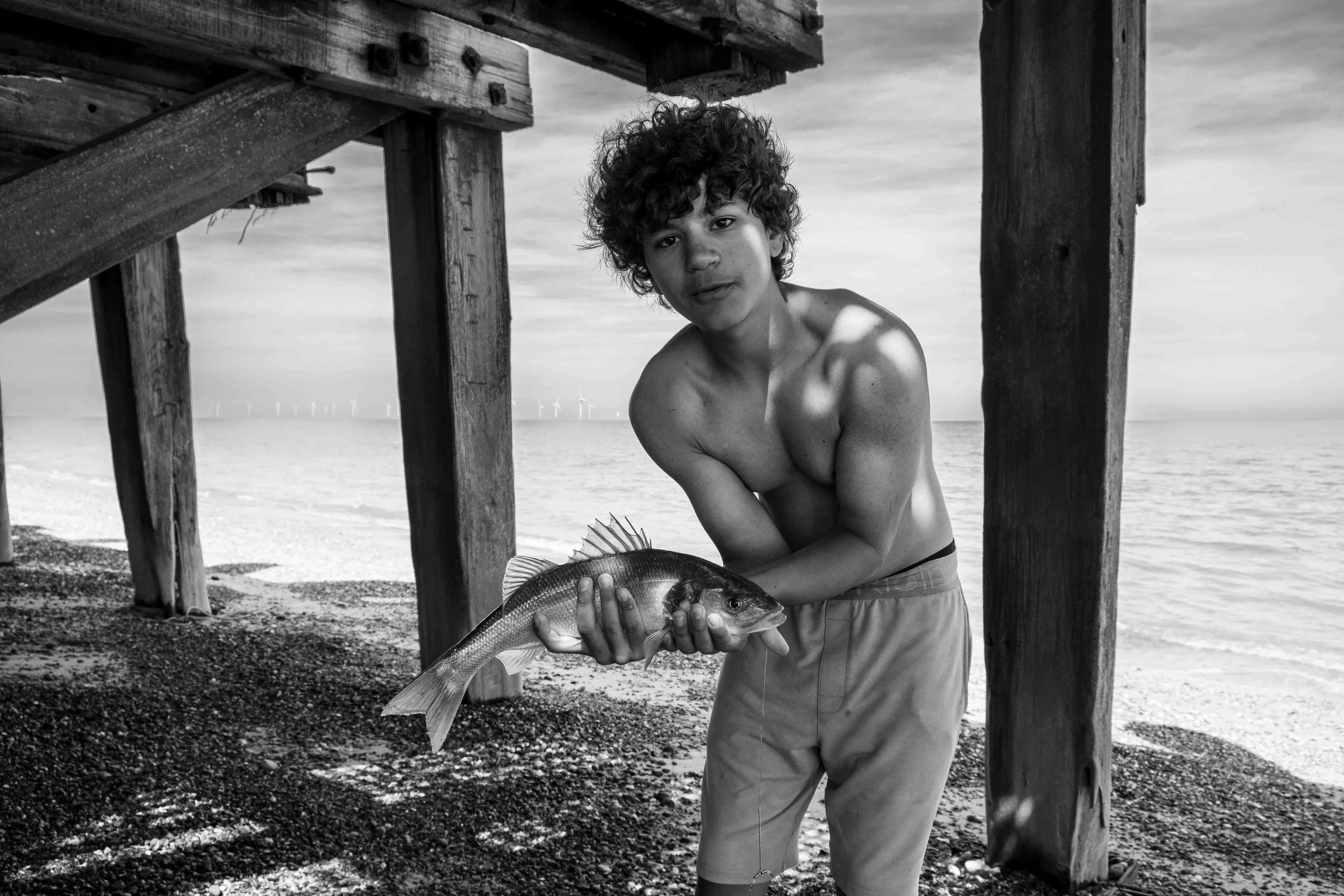 A young man with curly hair, shirtless, stands under a wooden pier on a beach, holding a fish he caught, with ocean waters and wind turbines visible in the background.