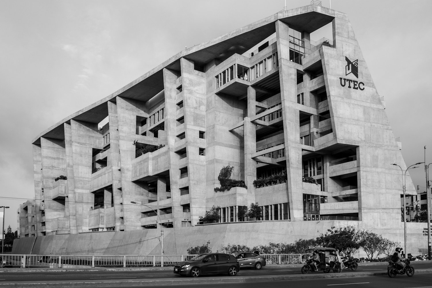 A modern architectural building made of concrete with multiple open balconies, and the logo UTEC on the side, situated by a street with cars, motorcycles, and scooters passing by.