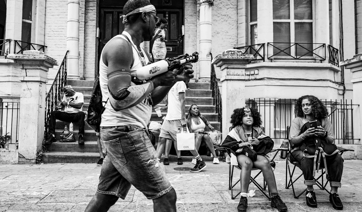 A street scene in black and white shows a man walking with a water gun, two women sitting on camping chairs, and other people on stairs and the sidewalk outside a building with a door and windows.