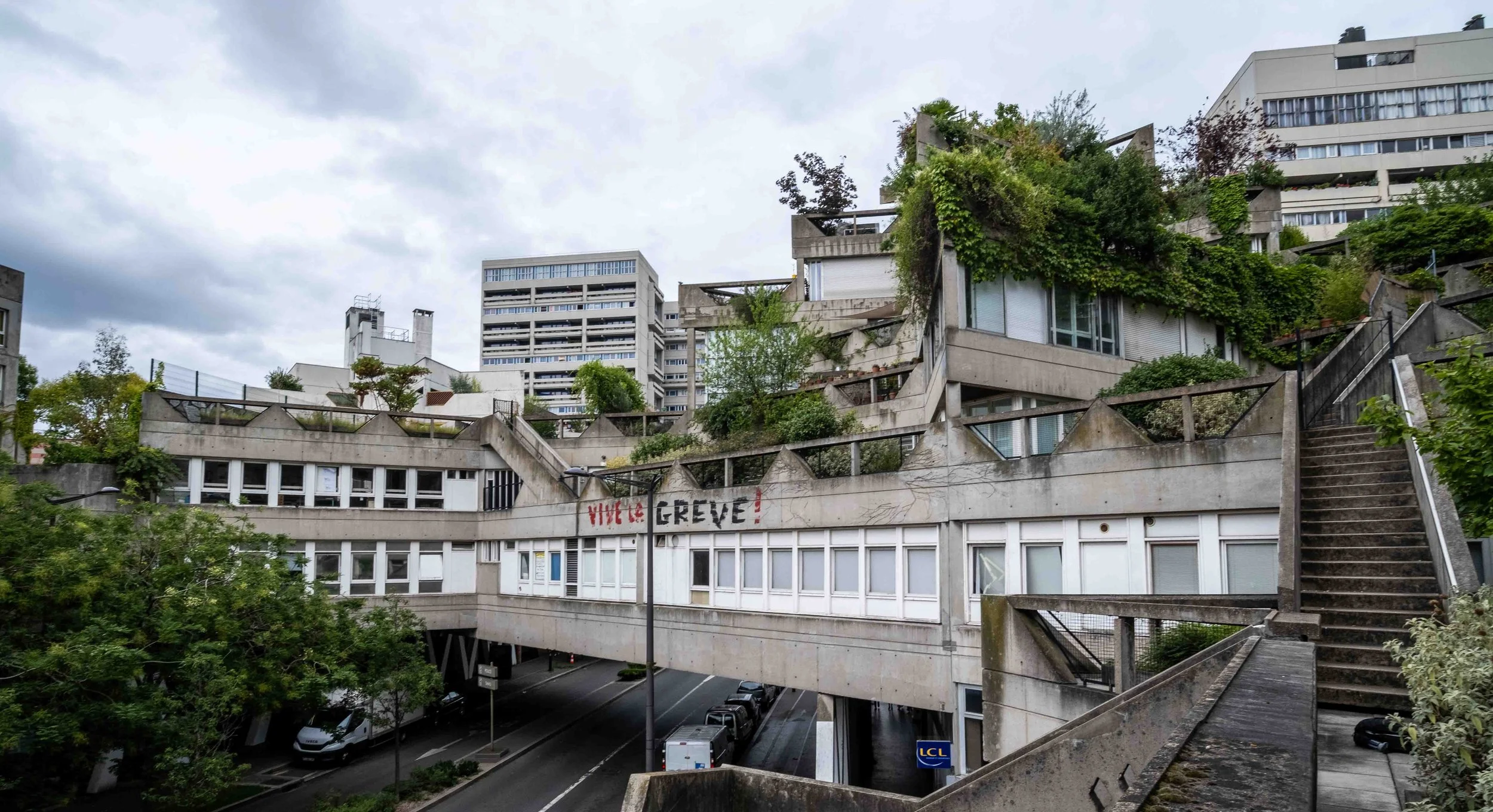 View of a modern Brutalist apartment complex with multiple levels and terraces covered in greenery, concrete stairs, and cars parked below. Some graffiti with red lettering reads 'VIVE LA GREVE!'