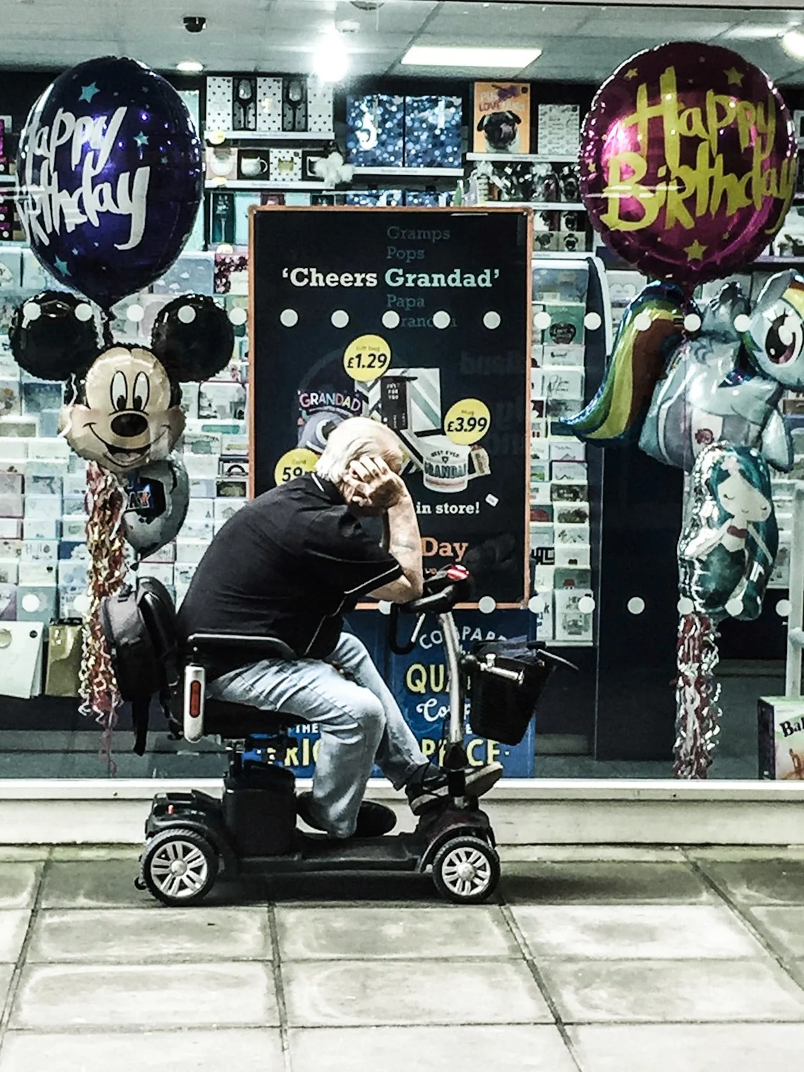An elderly man sitting on a mobility scooter outside a store window decorated with birthday balloons, including Mickey Mouse and unicorn themed balloons, during daytime.