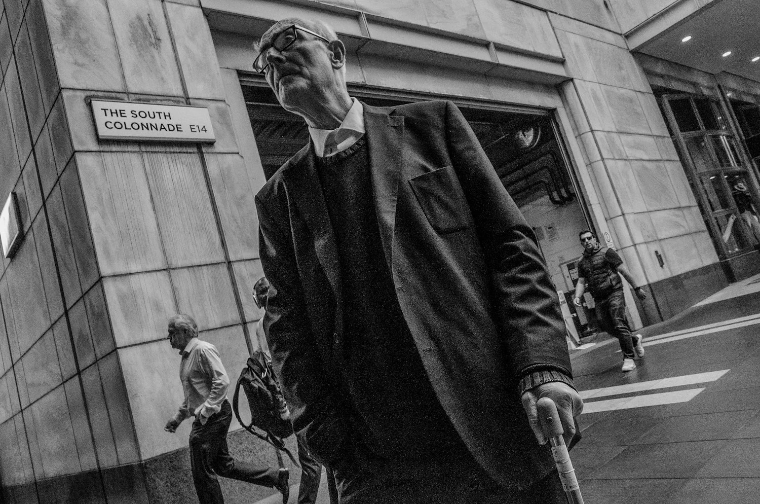 Black and white photo of an elderly man walking outside a building with a sign that reads "The South Colonnade E14." The man is wearing glasses, a suit, and using a cane. Other pedestrians are visible in the background.